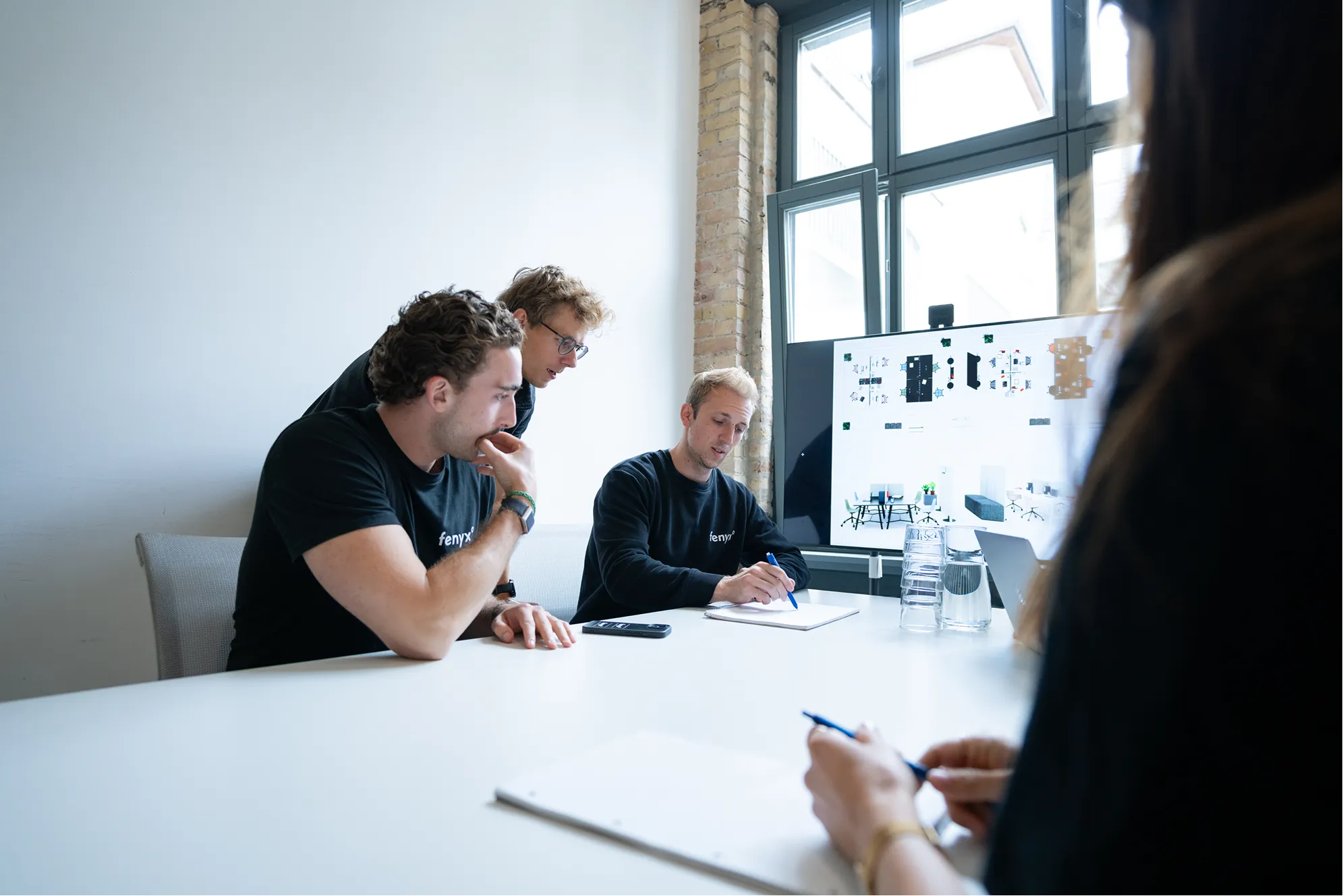 Three men and one woman in a meeting room discussing in front of a monitor displaying office layout designs.