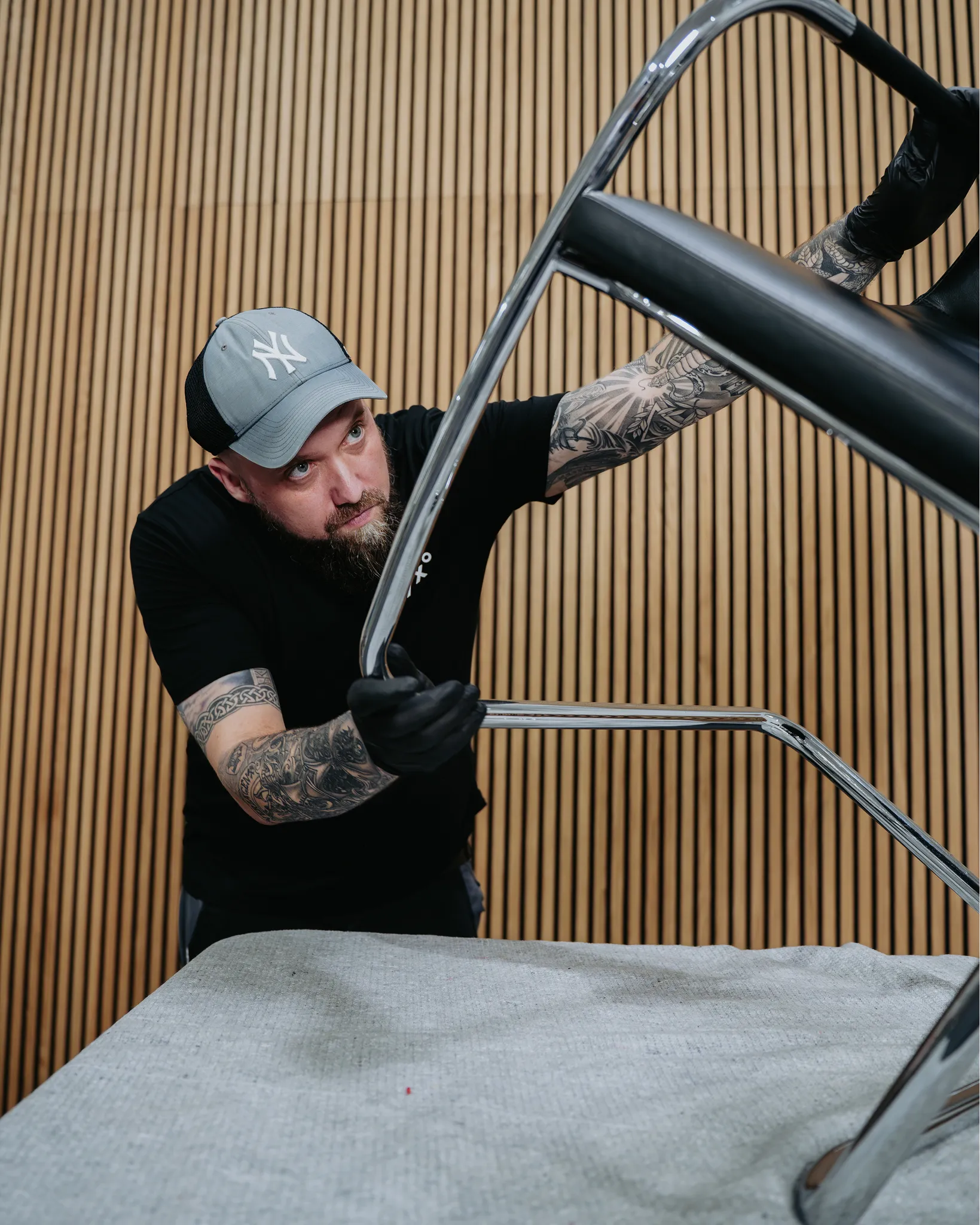 Man wearing a gray cap and black gloves inspecting the chrome frame of an upside-down chair in front of a wooden slat wall.