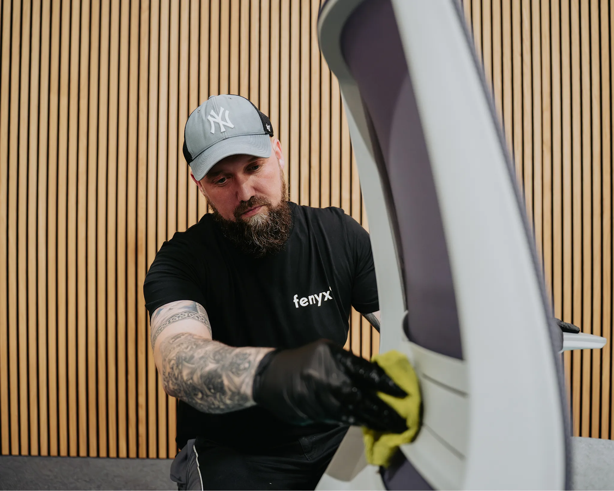 Man with tattoos wearing black gloves and a cap cleaning a white chair with a yellow cloth in front of a wooden slat wall.