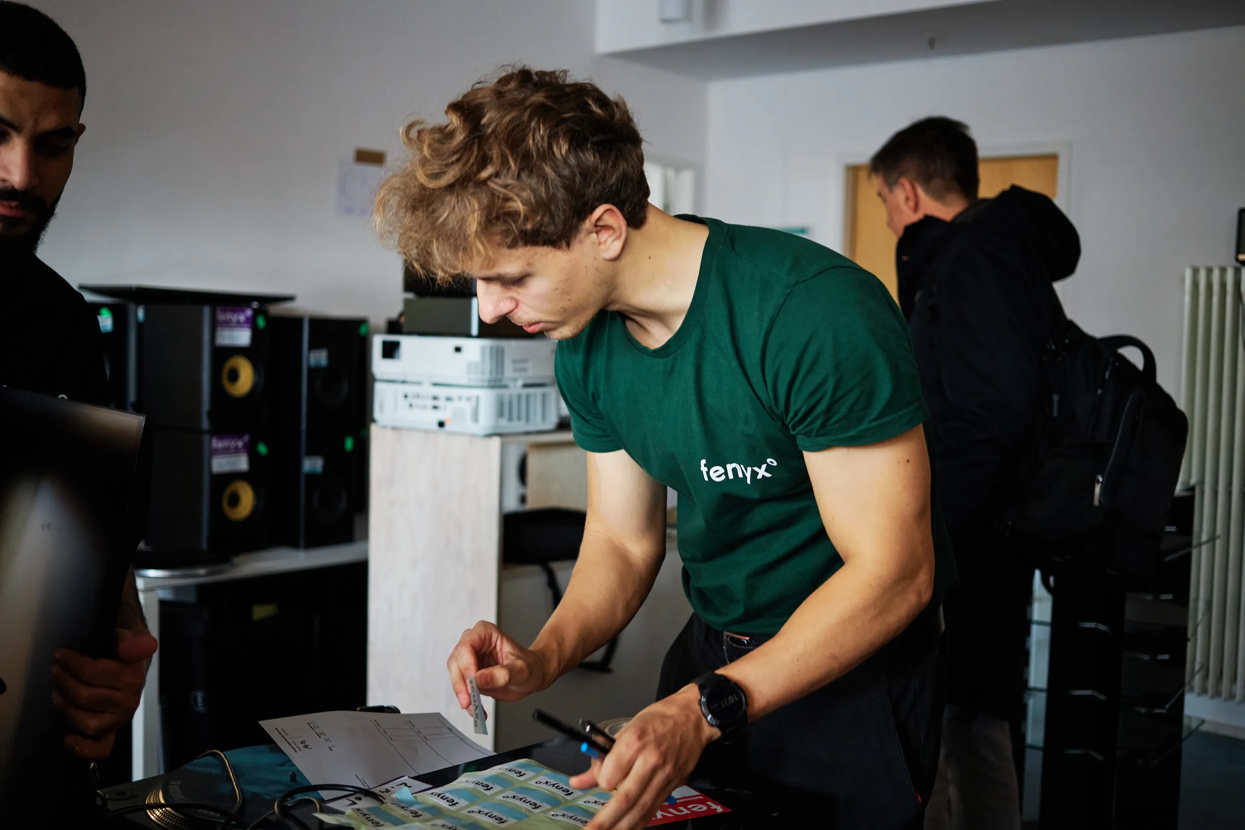 Young man in a green t-shirt with the text 'fenyx' leaning over a table with stickers and papers, interacting with another person.