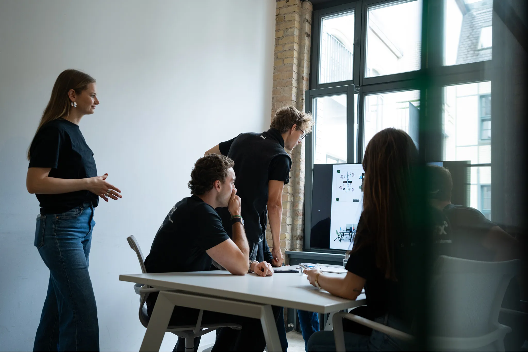 Four young professionals collaborating around a table, reviewing a floor plan displayed on a monitor in a modern office with large windows.