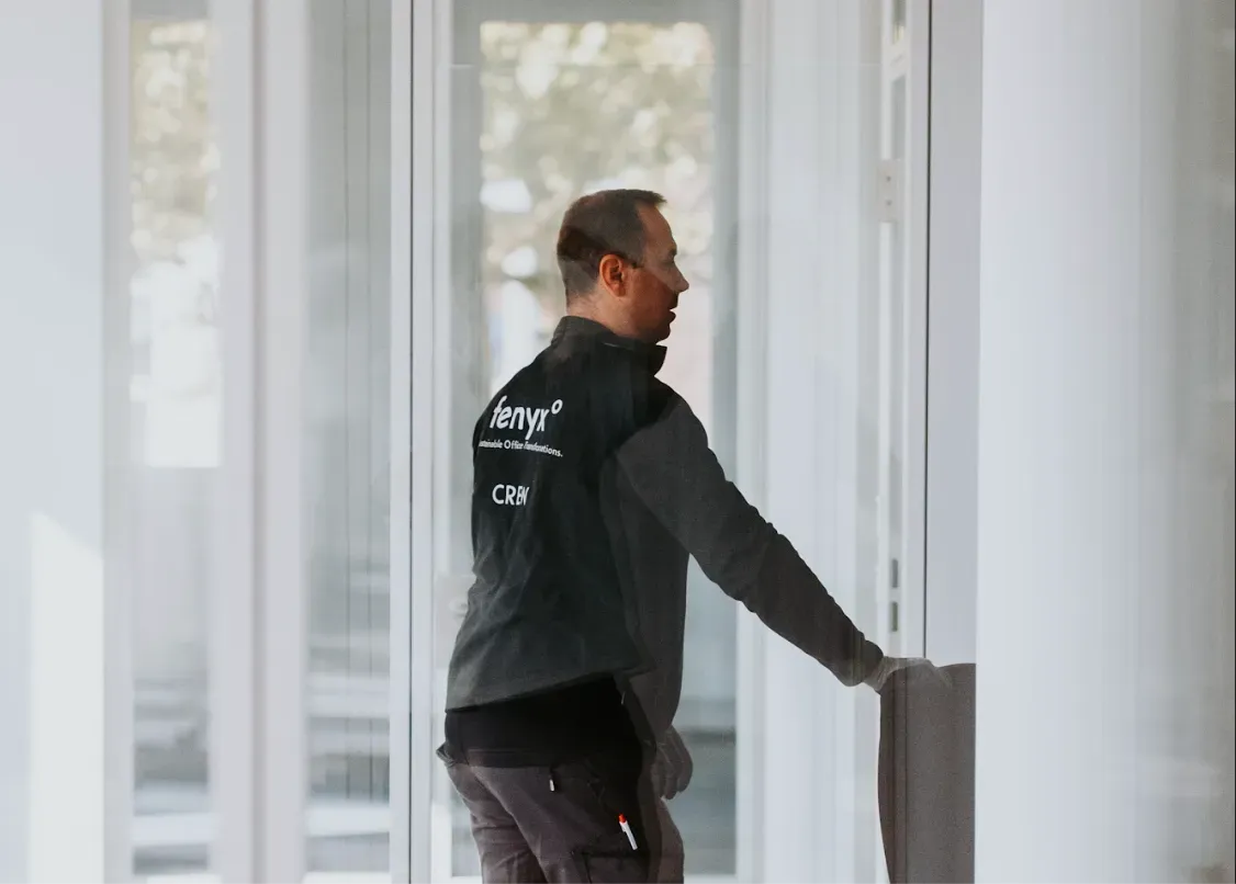 Man wearing a black jacket with text on the back, reaching out to open or touch a door in a bright, modern interior.
