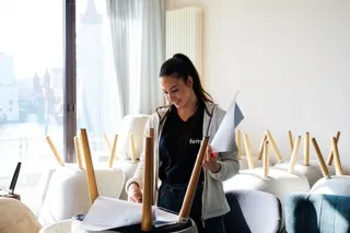 Young woman smiling while holding papers in a bright room with chairs stacked on tables.