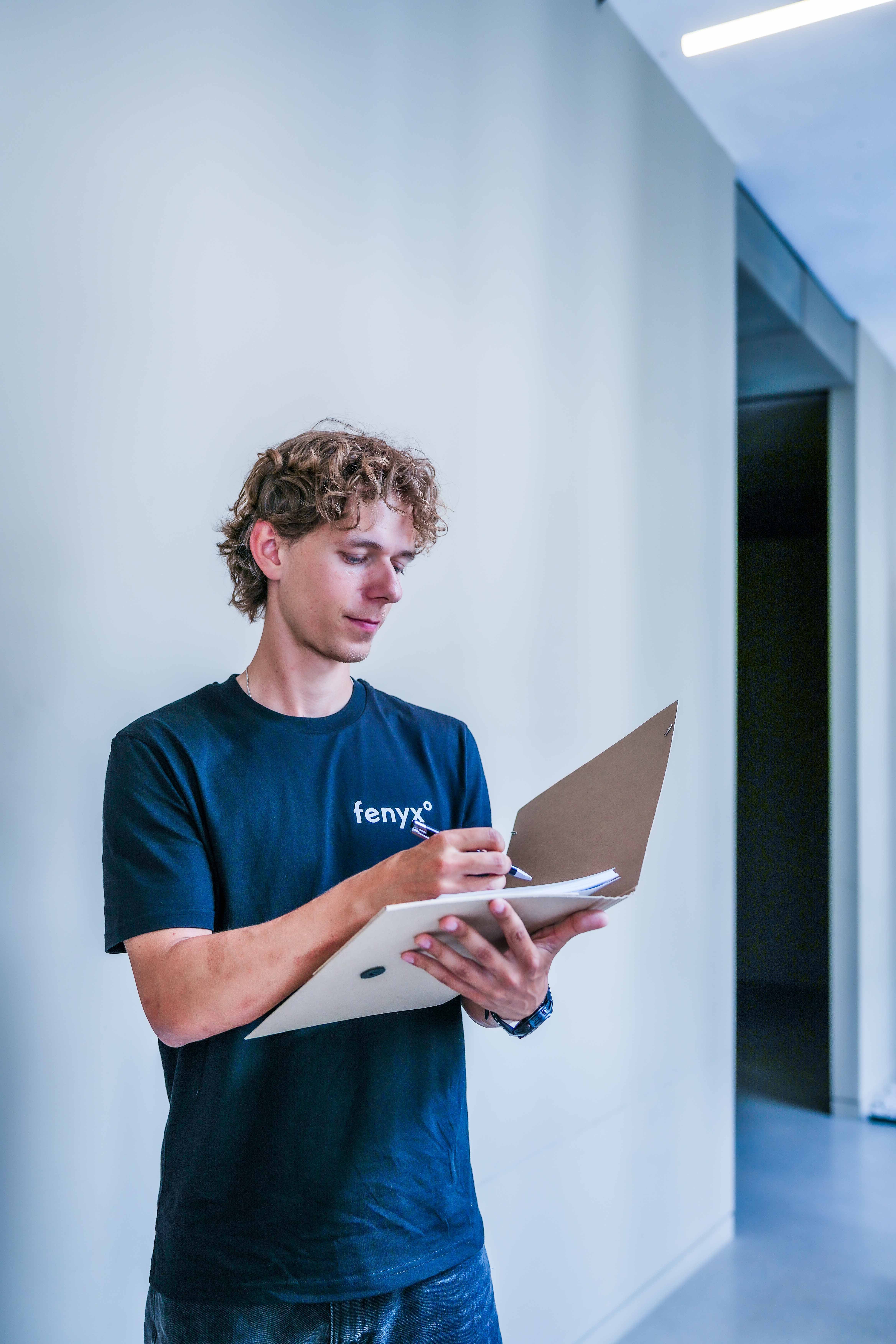 Young man with curly hair wearing a black Fenyx t-shirt writing on a clipboard in a hallway.