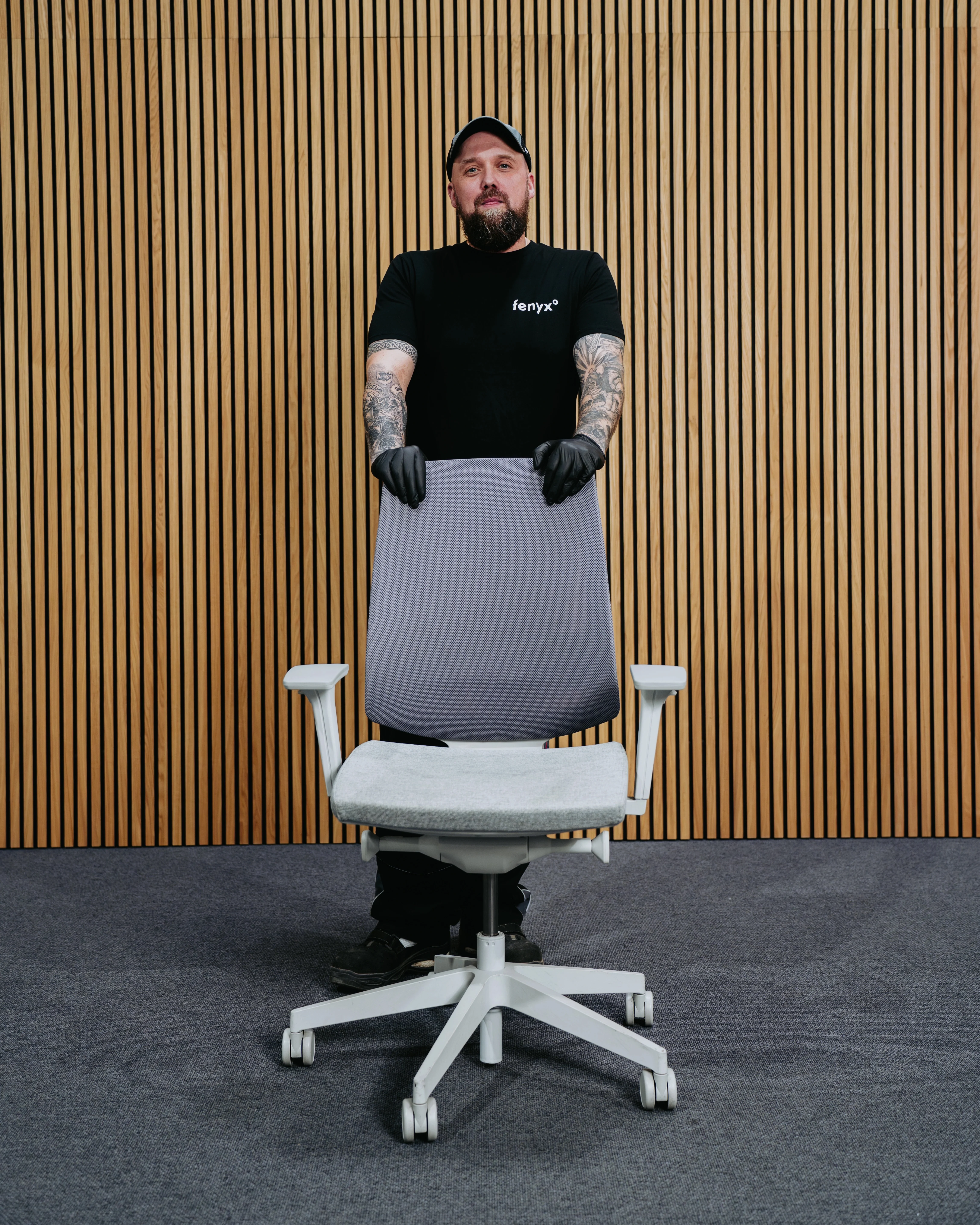 Man with tattoos wearing black gloves and a black shirt standing behind a gray office chair with wooden panel wall background.