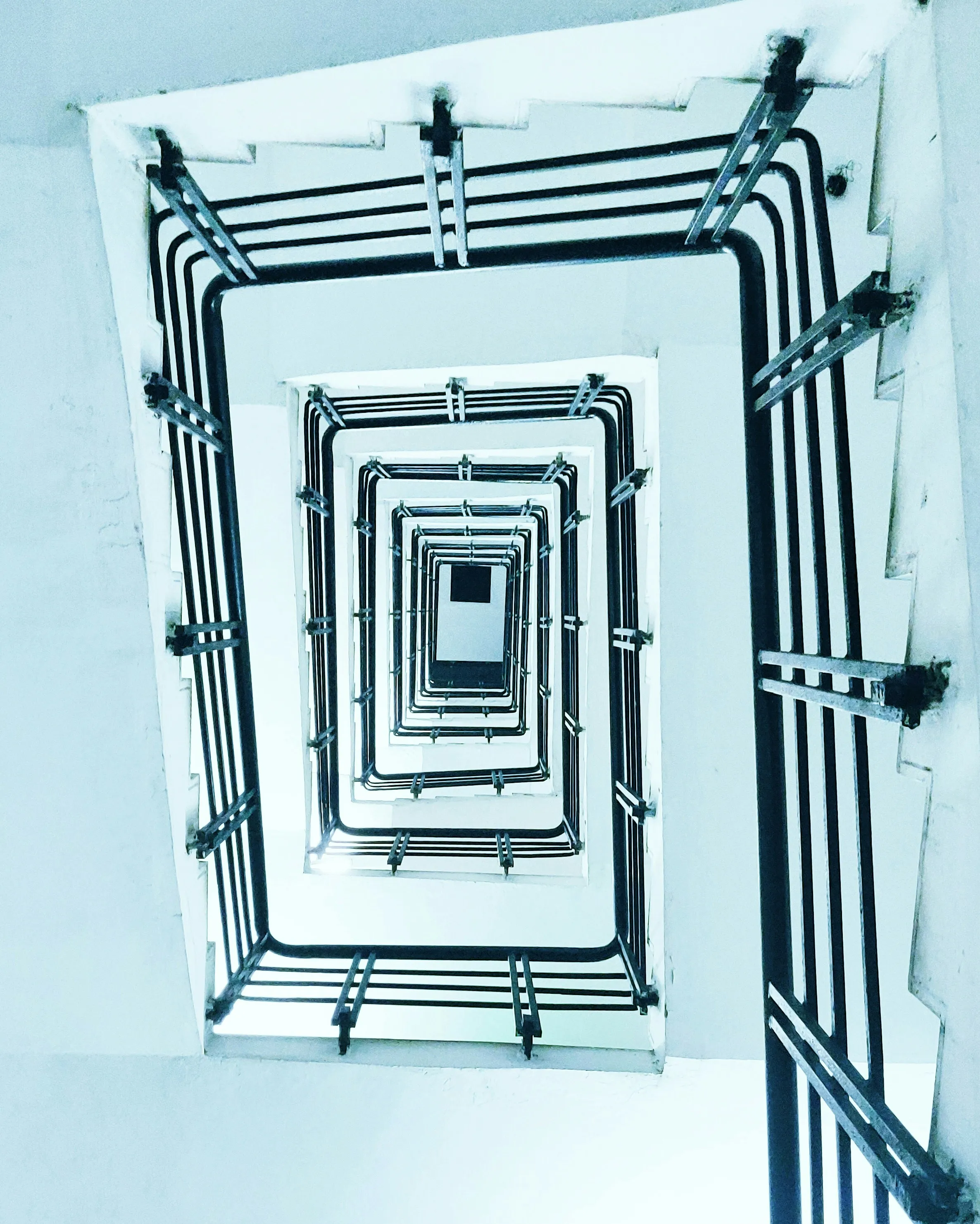 View looking up through a square spiral staircase with white walls and black railings.