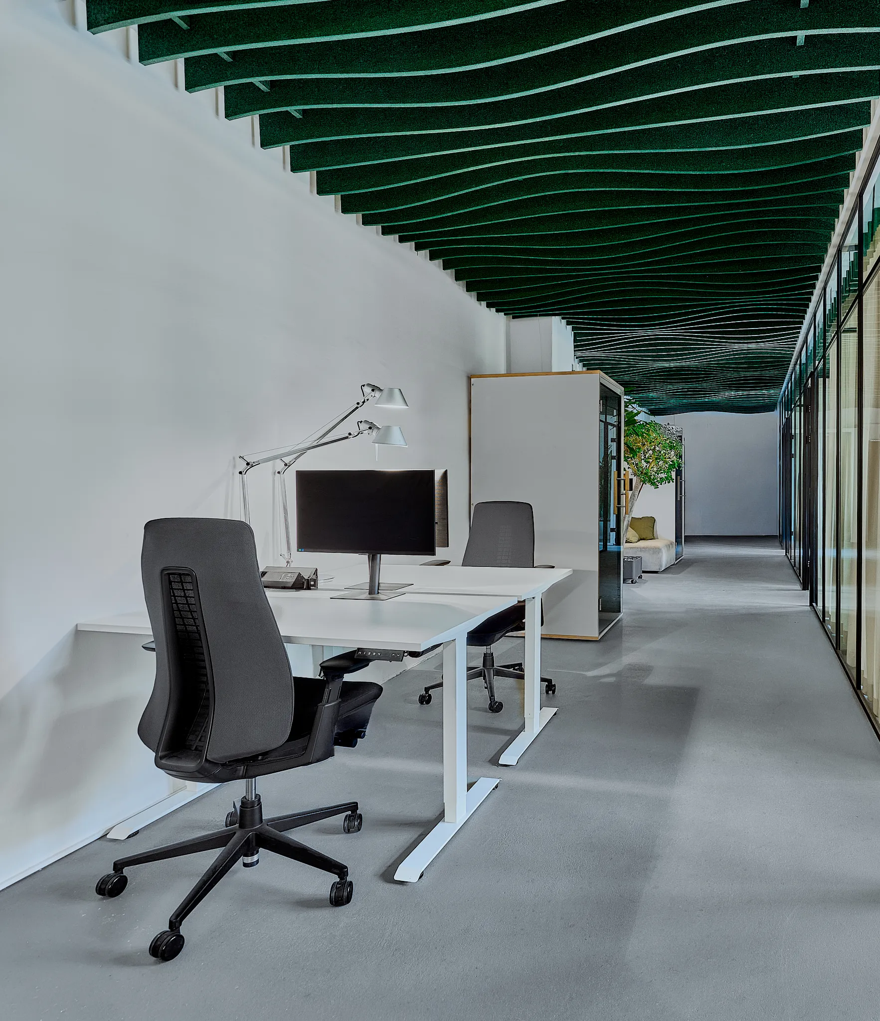 Modern office workspace with two black ergonomic chairs, a white desk, dual desk lamps, and a computer monitor under a ceiling with green sound-absorbing panels.