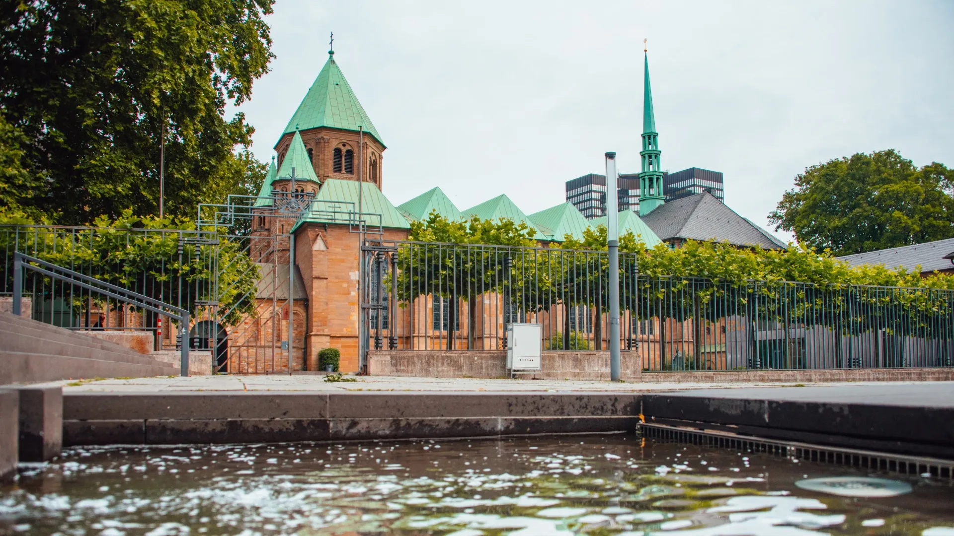 Historische Kirche mit kupfergrünen Dächern hinter einem Zaun und Wasserspiegel im Vordergrund.