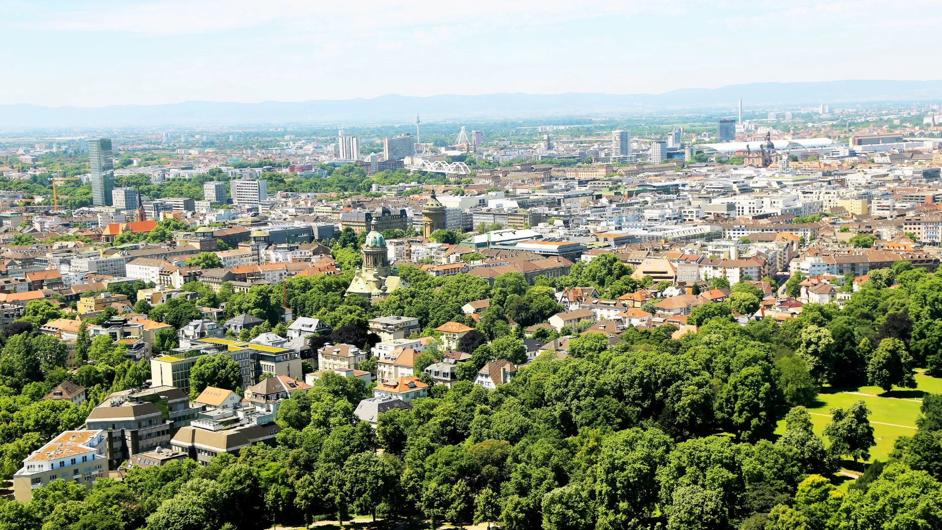 Panorama der Stadt Mannheim mit grünen Parks, Gebäuden und Hügeln im Hintergrund.