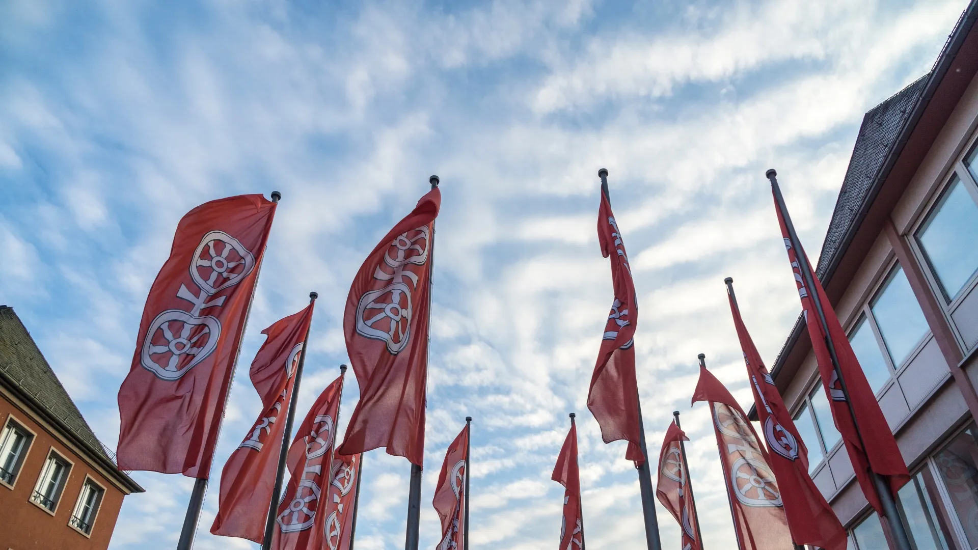 Mehrere rote Flaggen mit weißem Radsymbol wehen vor Himmel und Gebäuden in Mainz.