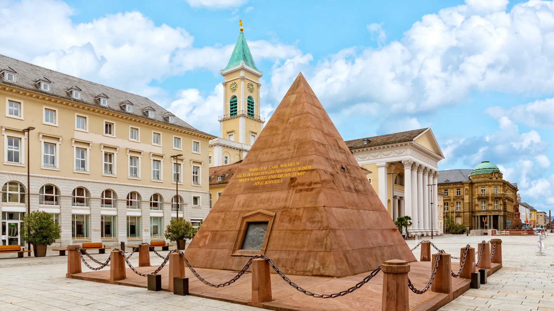 Sandsteinpyramide auf dem Schlossplatz in Karlsruhe mit historischen Gebäuden und blauem Himmel.