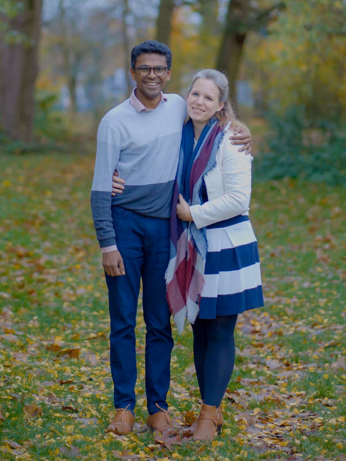 Smiling and embracing while standing on a leaf-covered grassy path.