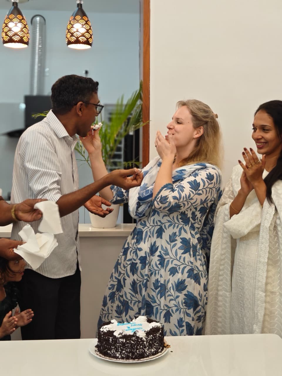 Celebrating with cake, Alexandra feeds cake to Jithu while others clap and smile nearby.