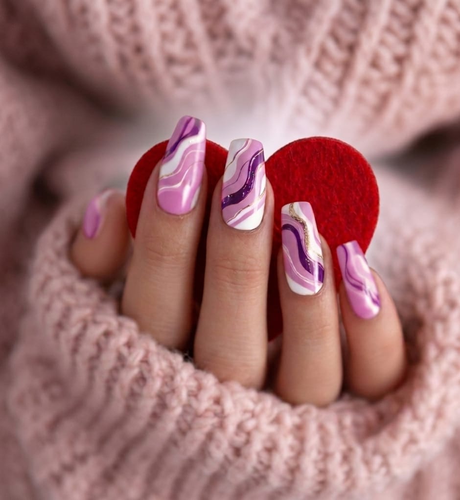 Hand met roze en paarse golvende nagelkunst, houd rood vilten hart vast, omgeven door een zachte roze trui.