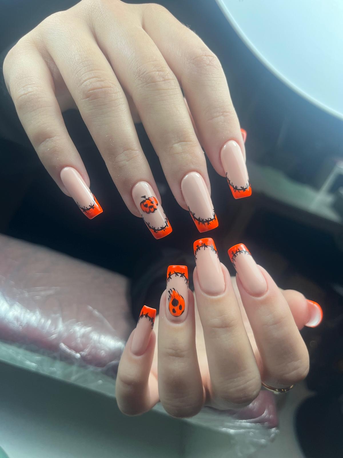 Hand met lange nude nagels met oranje franjes en zwarte spijkerontwerpen, inclusief pompoen- en vlammendetail voor Halloween.