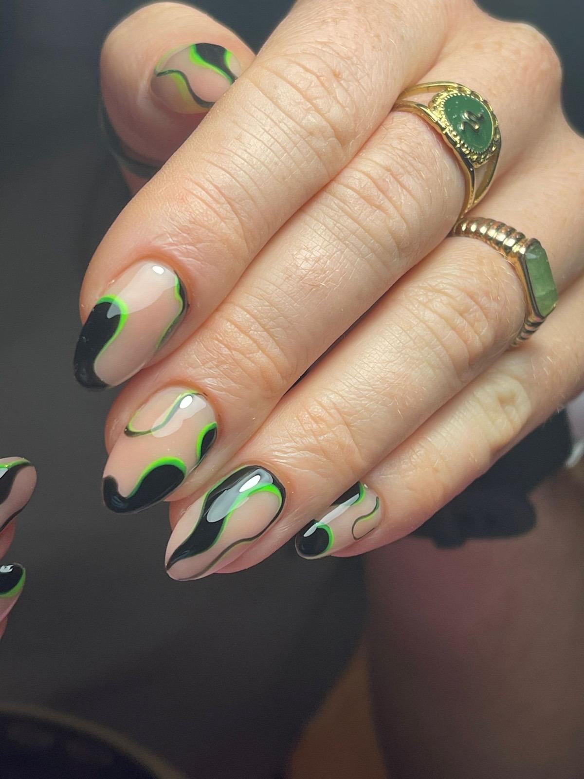 Hand met nagels in glanzende nude kleur met zwart-groene abstracte golvende lijnen en gouden ringen met groene stenen.