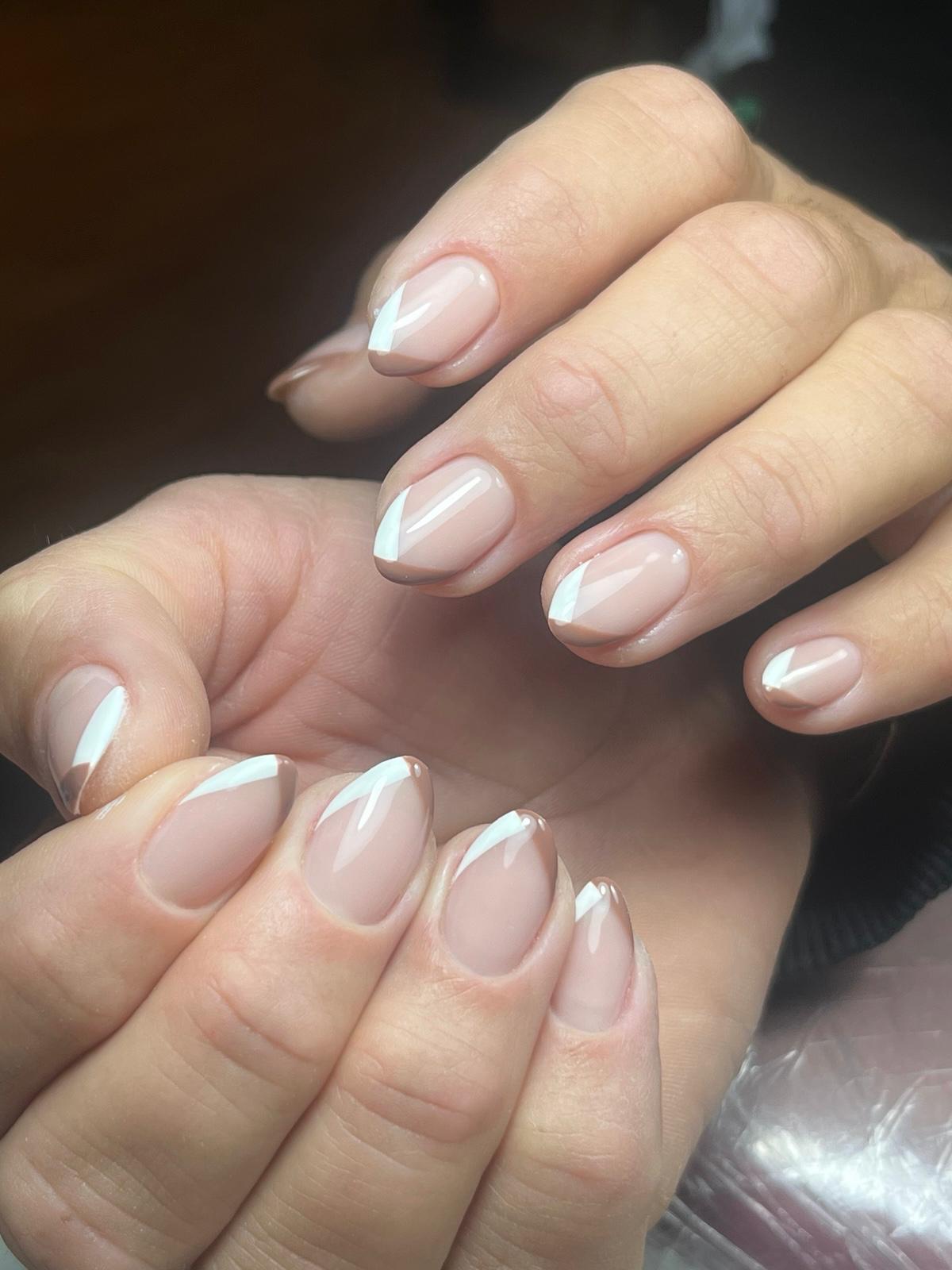 Handen met korte, ovale nagels gelakt in zacht roze met een diagonale witte en bruine Franse manicure.