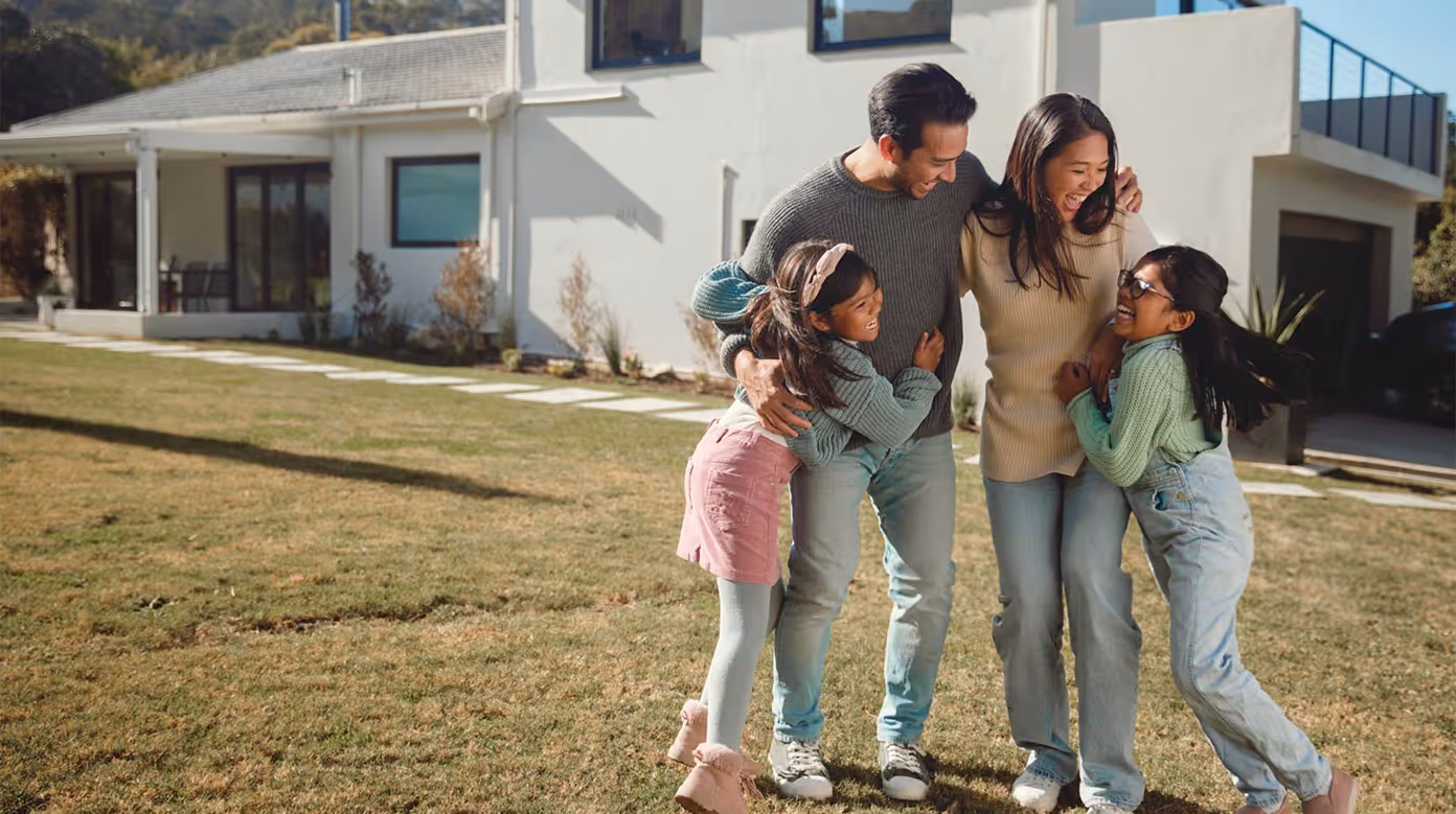 Happy family of four hugging and laughing together outside their modern house on a sunny day.