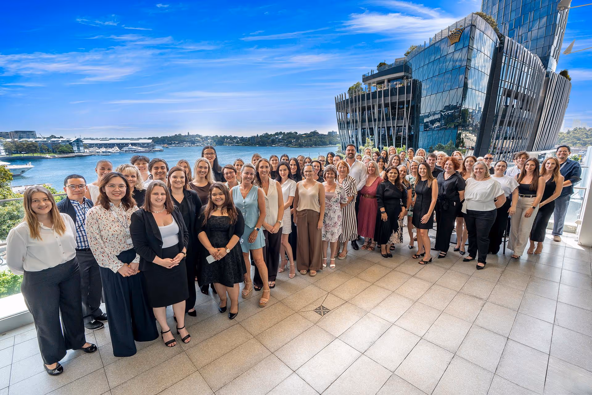 Group of diverse professionals posing on a terrace with water and a modern glass building in the background under a blue sky.