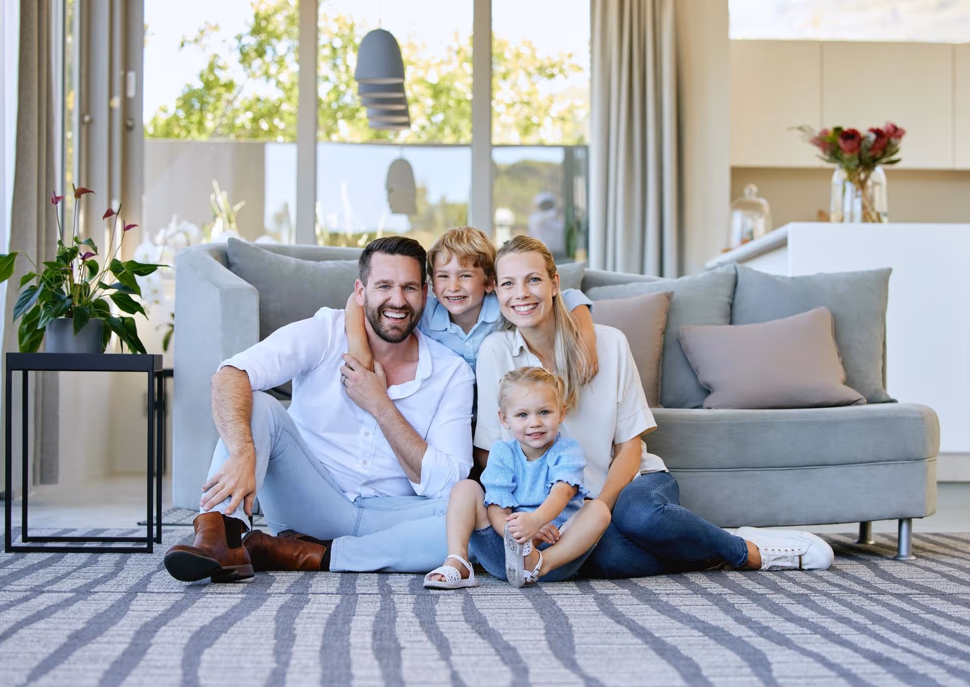 Smiling family of four sitting together on a carpeted floor in a bright living room with a grey sofa behind them.