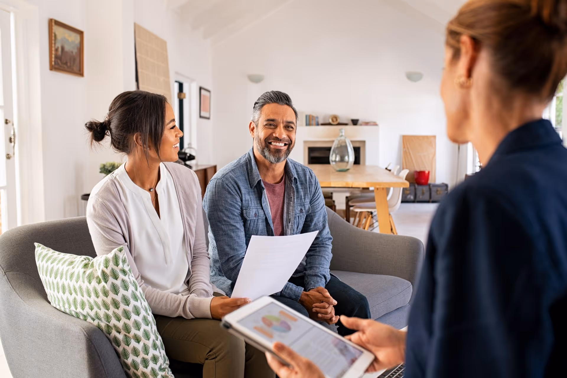 Smiling couple sitting on a couch holding a document while talking to a professional with a tablet in a bright living room.