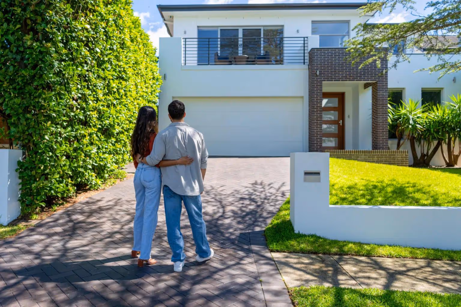 Couple standing arm in arm looking at modern two-story house with a green hedge and lawn.