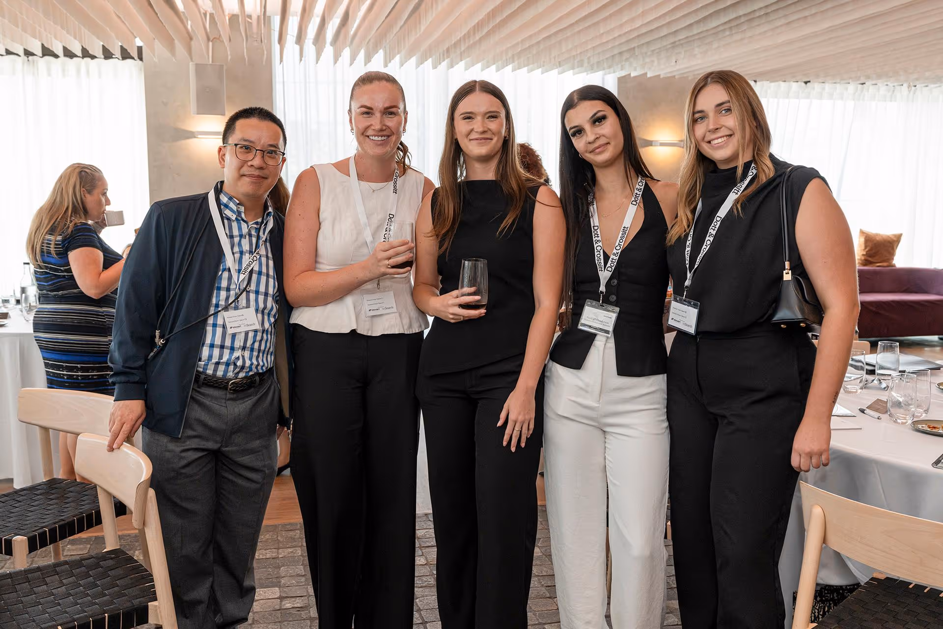 Group of five smiling conference attendees wearing name badges, two holding drinks, posing indoors.