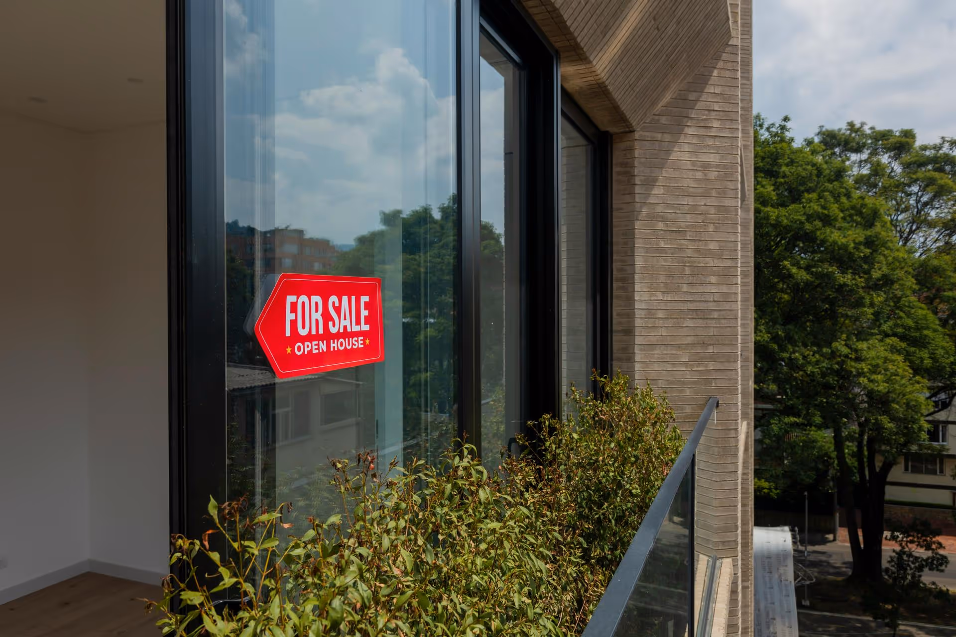 Red 'For Sale Open House' sign on the glass door of an empty apartment balcony with plants and trees outside.