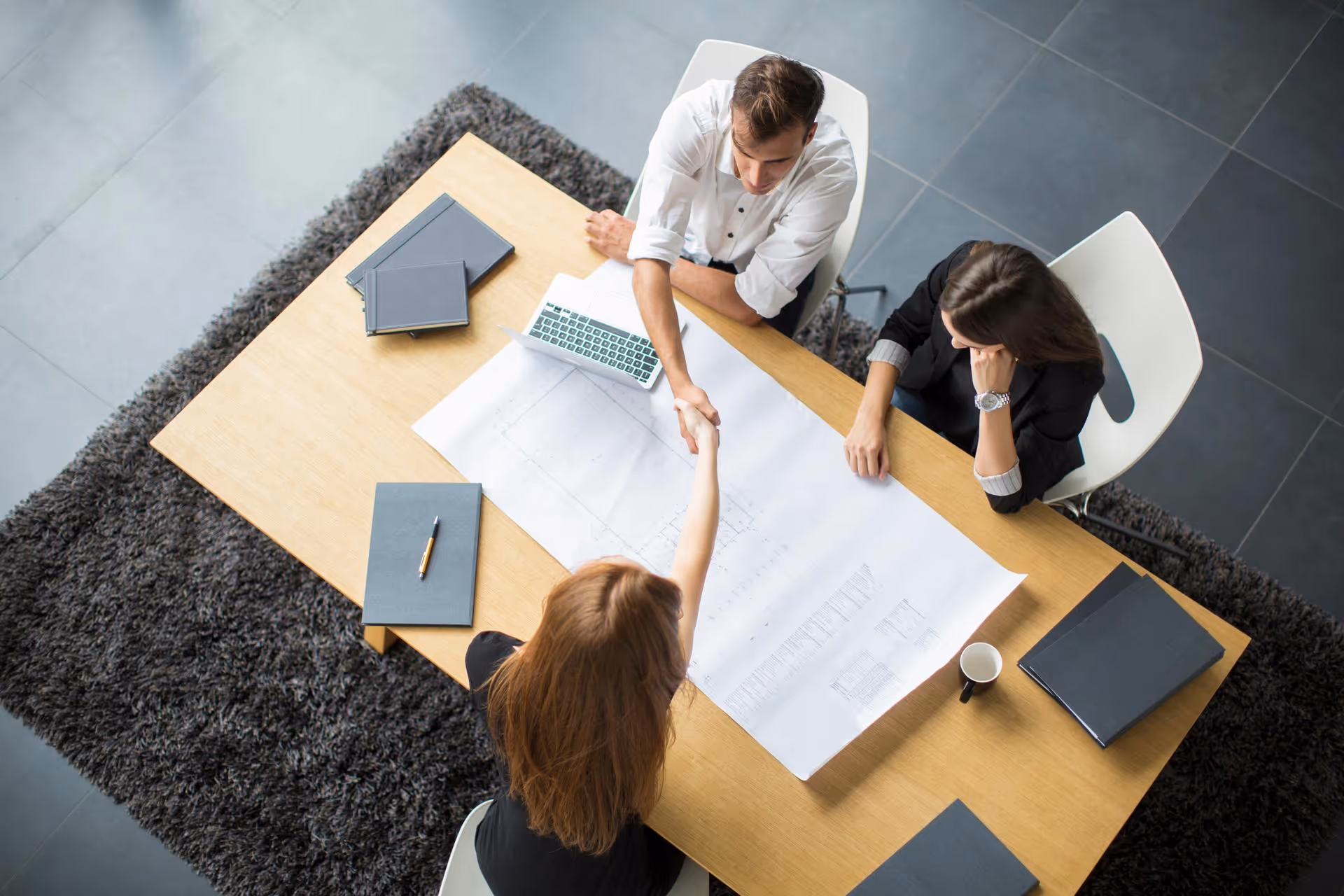 Two women and one man sitting at a table with architectural plans, shaking hands.