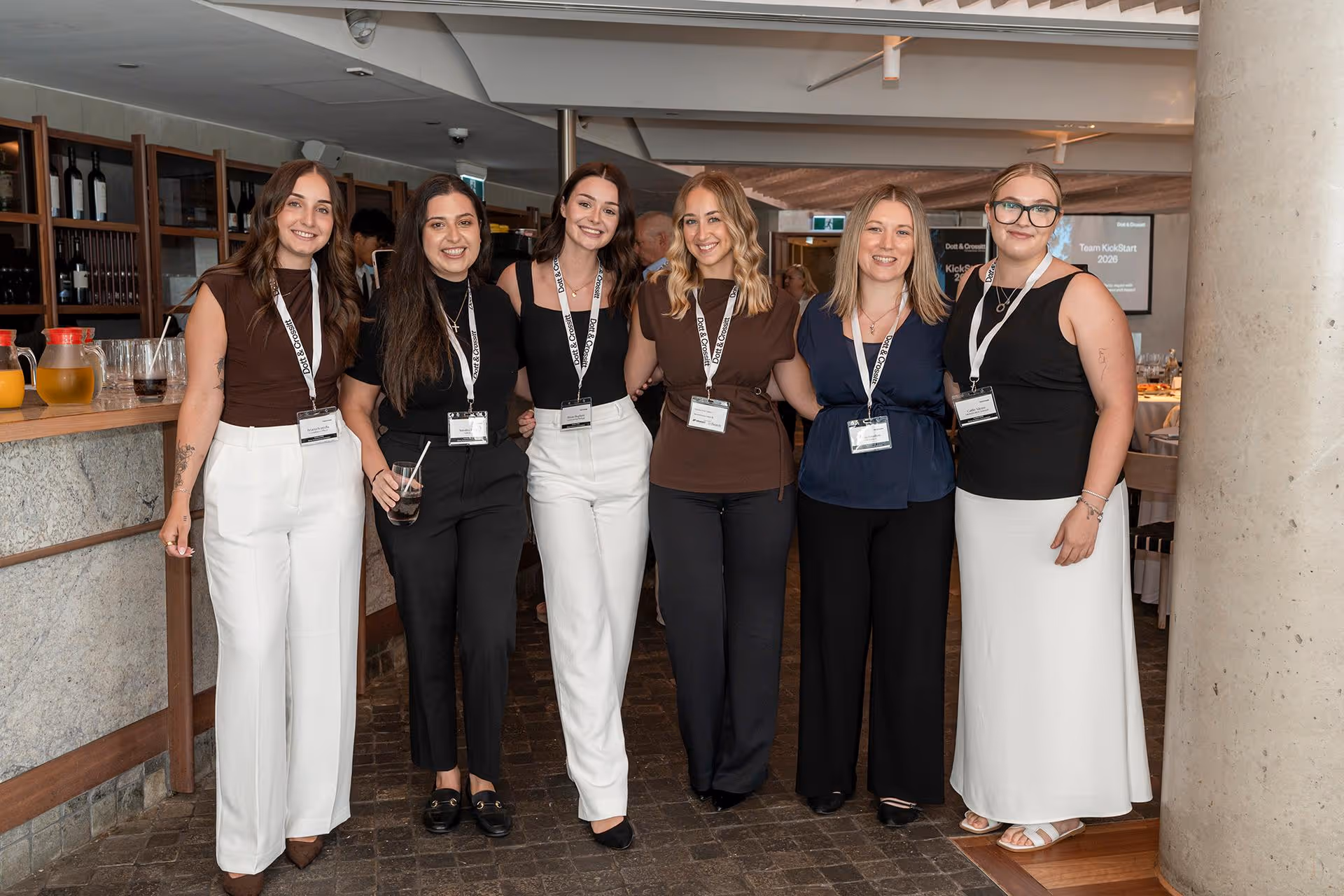 Six women standing side by side indoors, smiling, wearing name badges on lanyards at a social event.