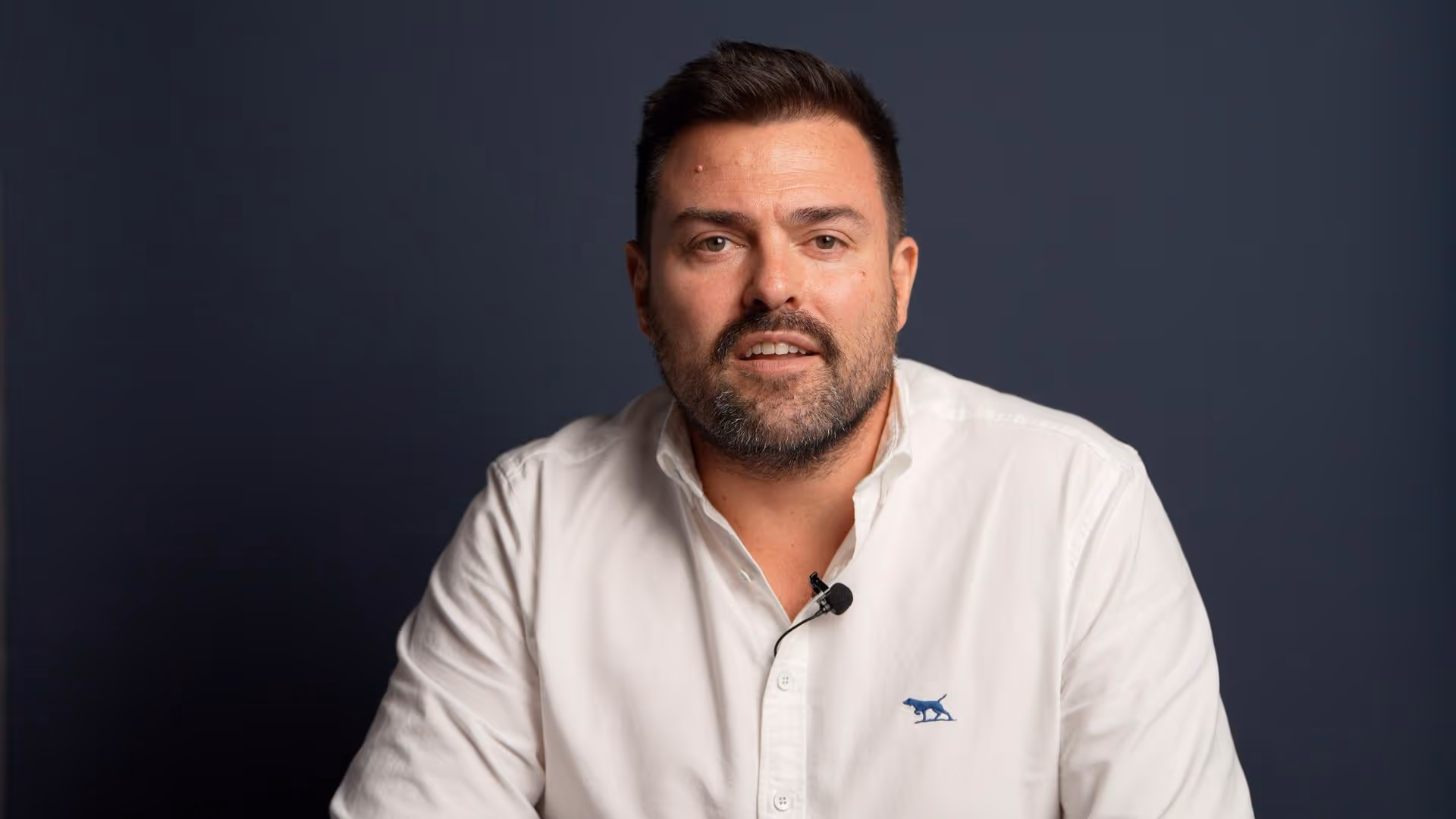Man with short dark hair and beard wearing a white button-up shirt with a blue panther logo, speaking in front of a dark grey background.