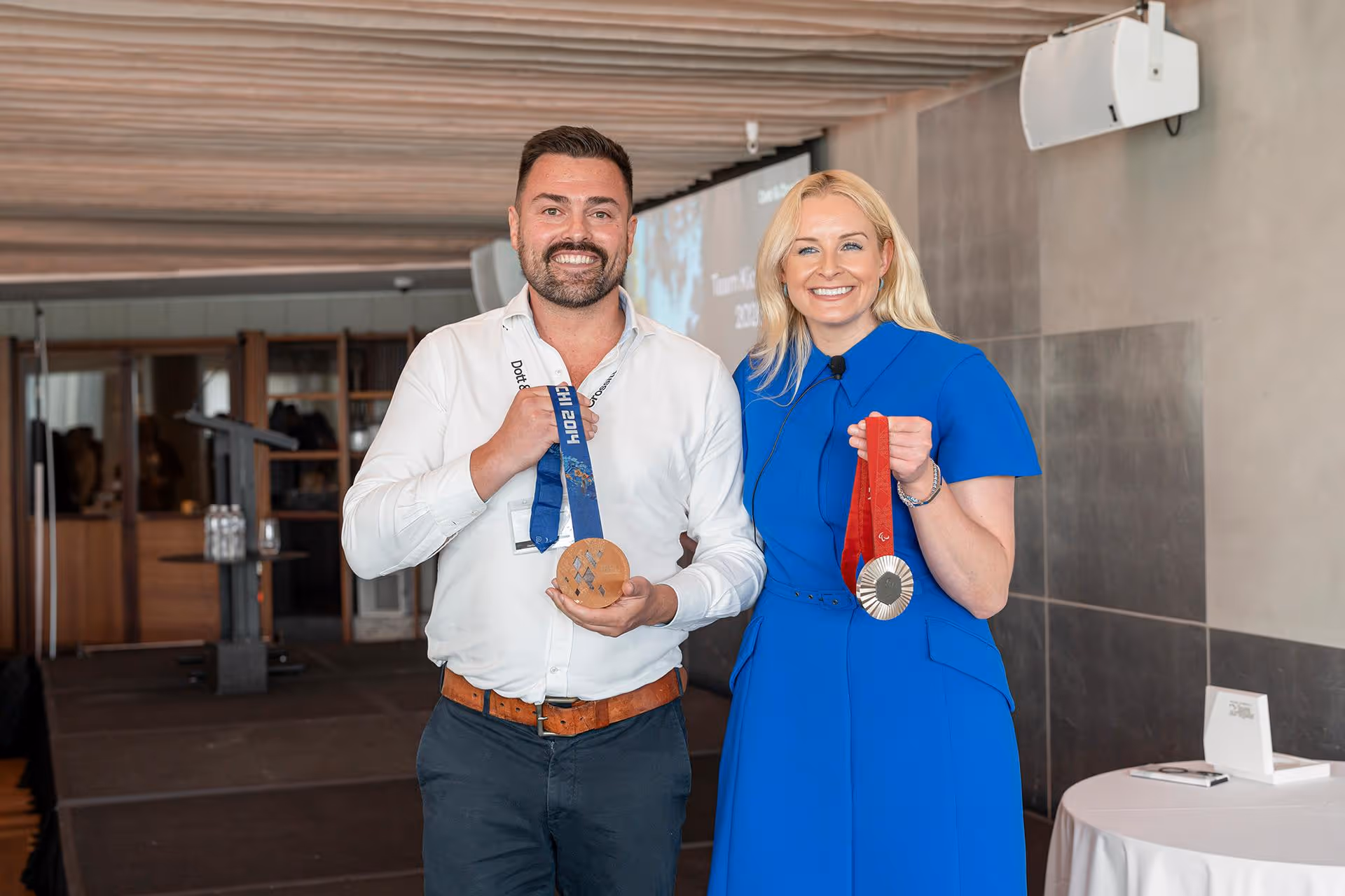 Smiling man and woman standing indoors holding medals, man has a bronze medal with a blue ribbon, woman has a silver medal with a red ribbon.