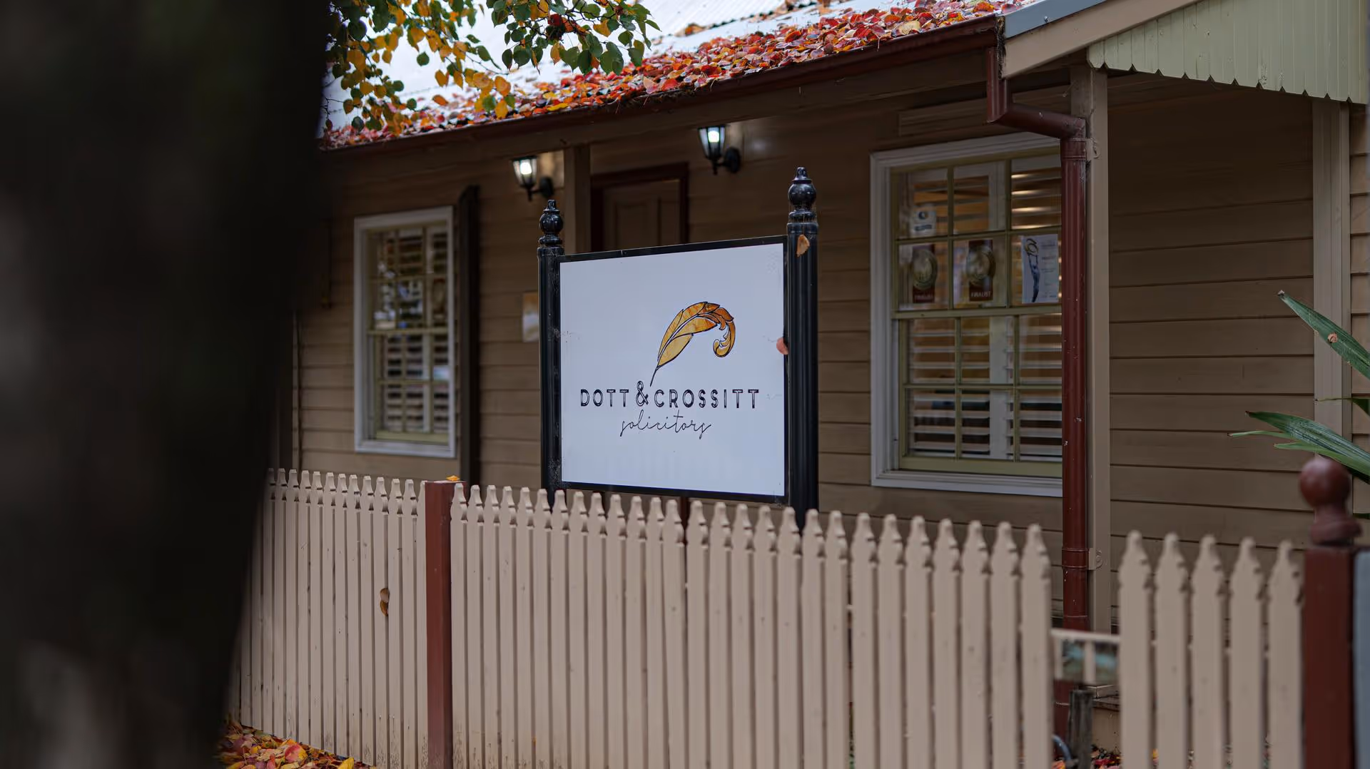 White sign with a feather logo and text reading 'Dott & Crossitt solicitors' outside a beige wooden building with white picket fence and autumn leaves on the roof.