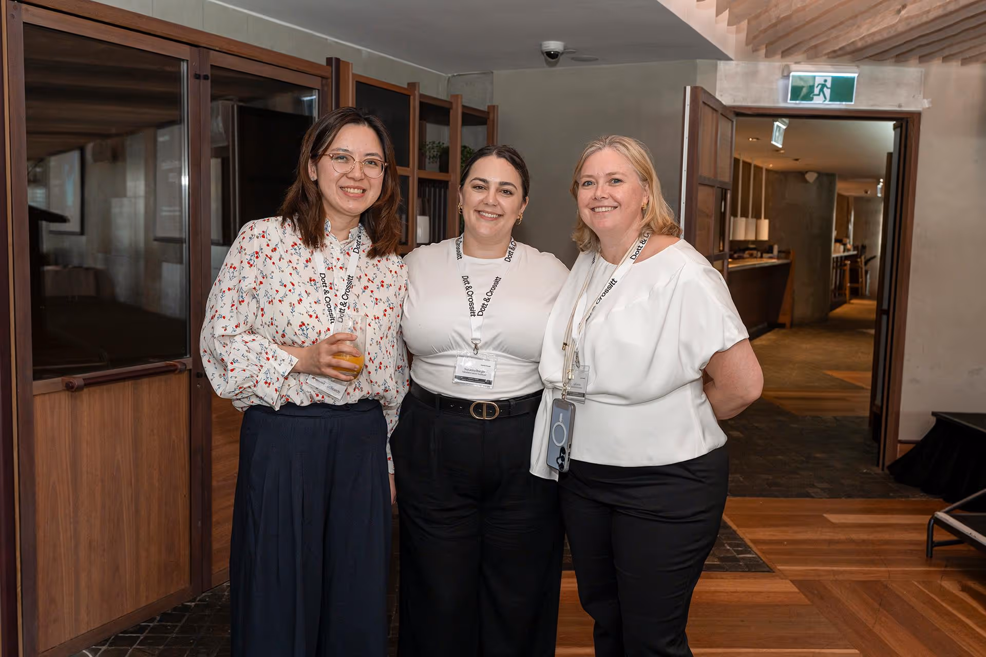 Three women smiling and posing indoors, wearing white tops and black pants, each with a conference lanyard and name badge.