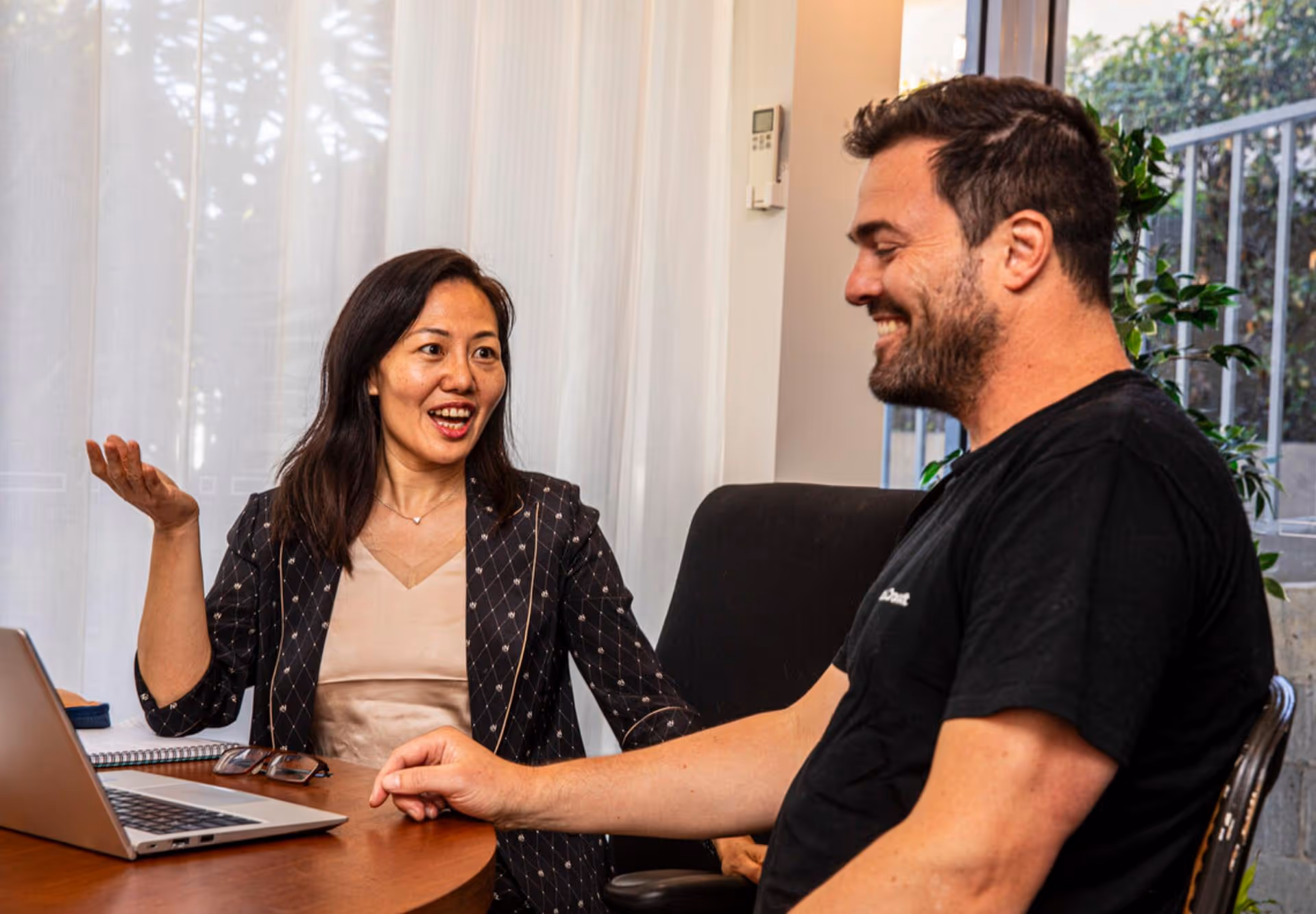 Two colleagues sitting at a table engaged in a cheerful discussion with a laptop and notebook in front of them.