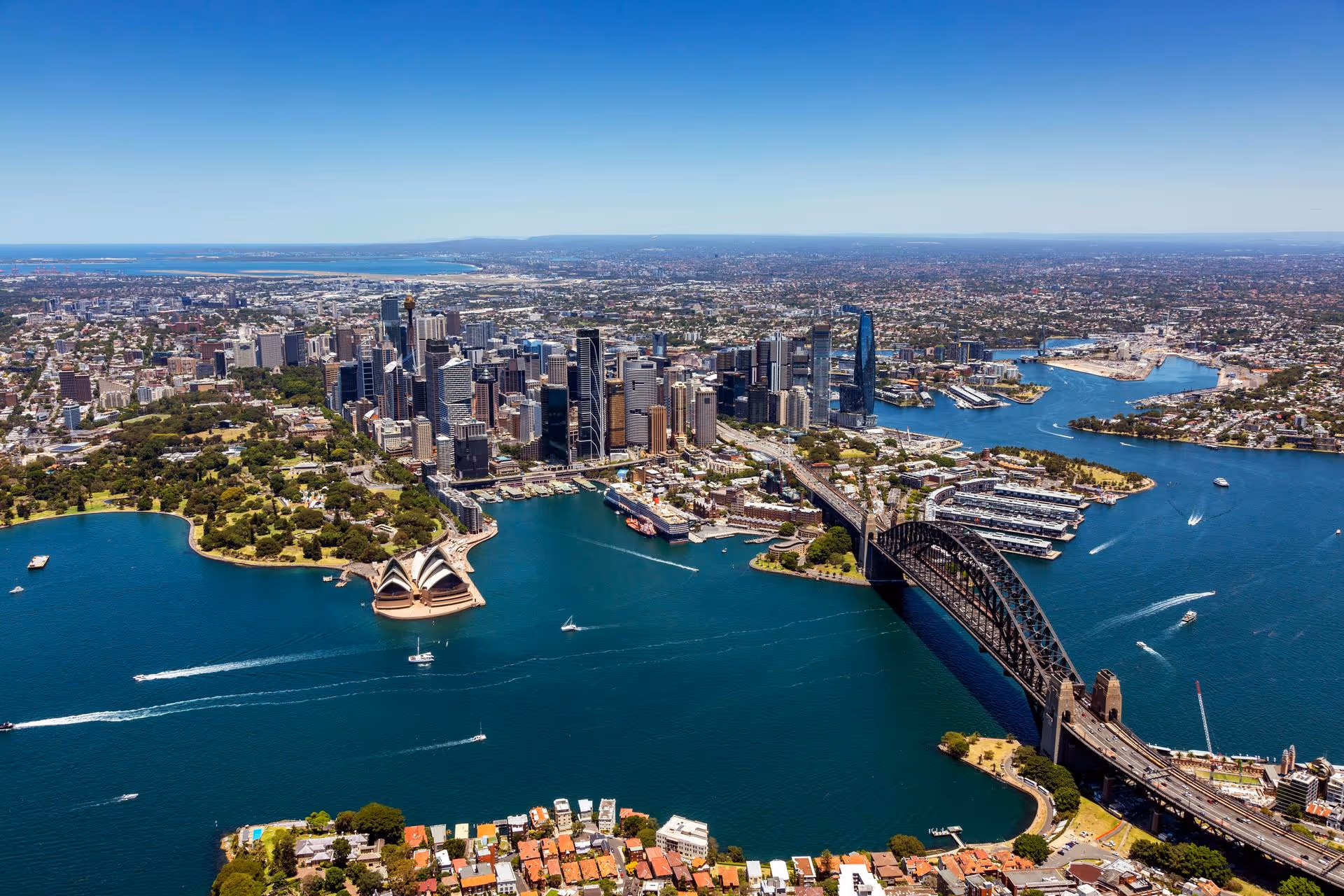 Aerial view of Sydney featuring the Opera House, Harbour Bridge, city skyline, and boats on the water.