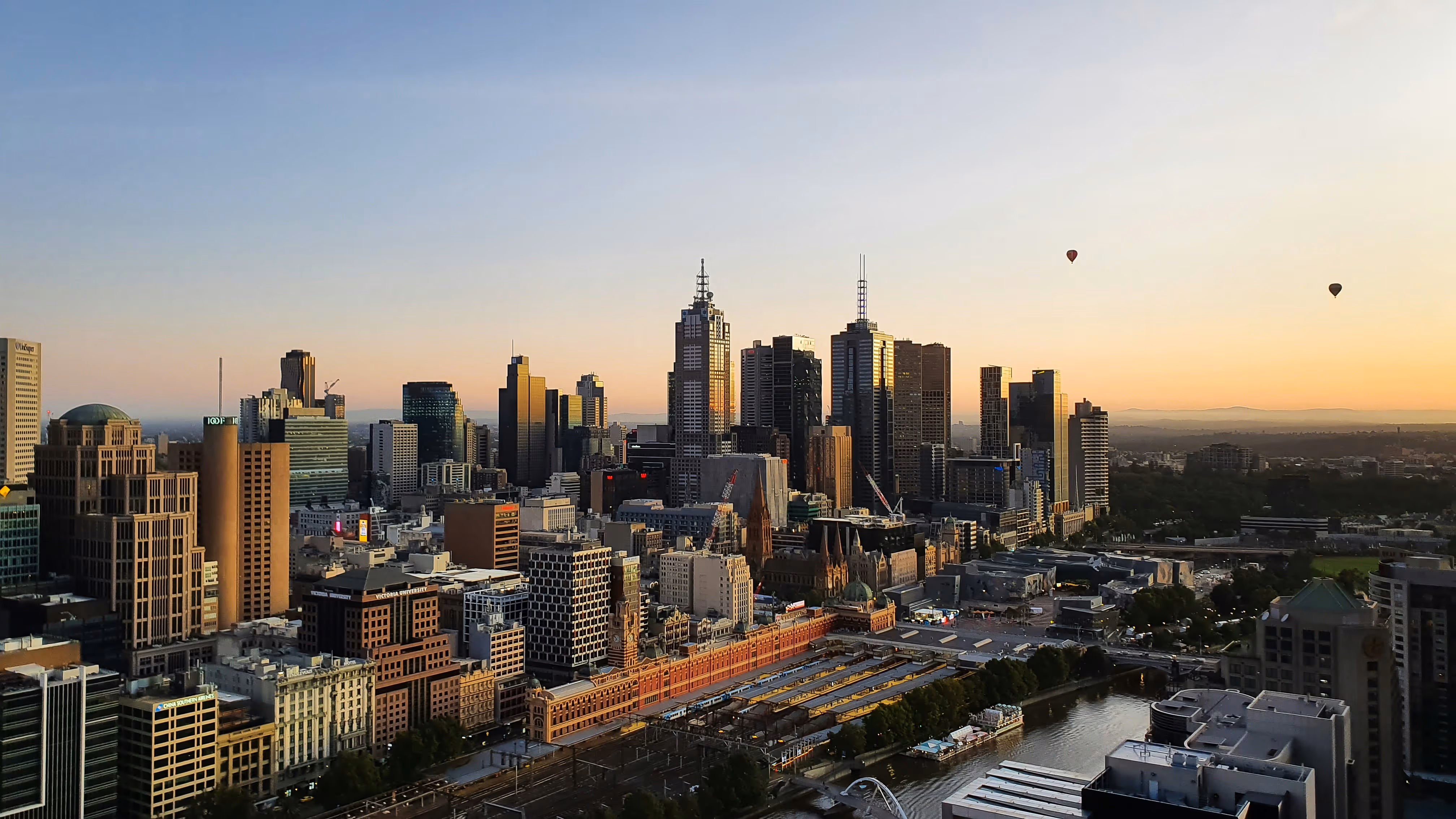 Melbourne city skyline at sunset with high-rise buildings, a river, and two hot air balloons in the sky.