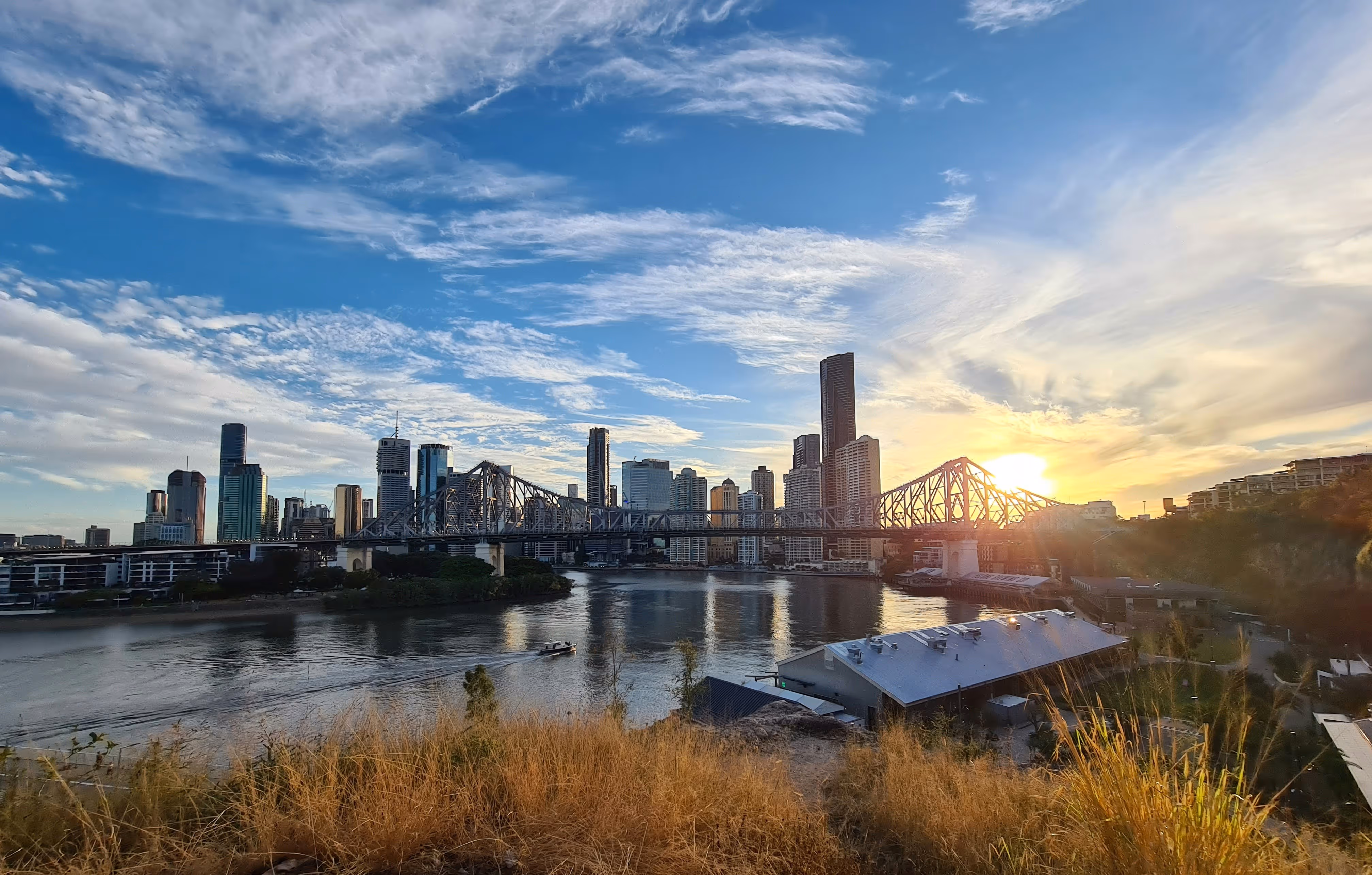 Brisbane city skyline at sunset with the Story Bridge over the Brisbane River and a boat cruising the water.