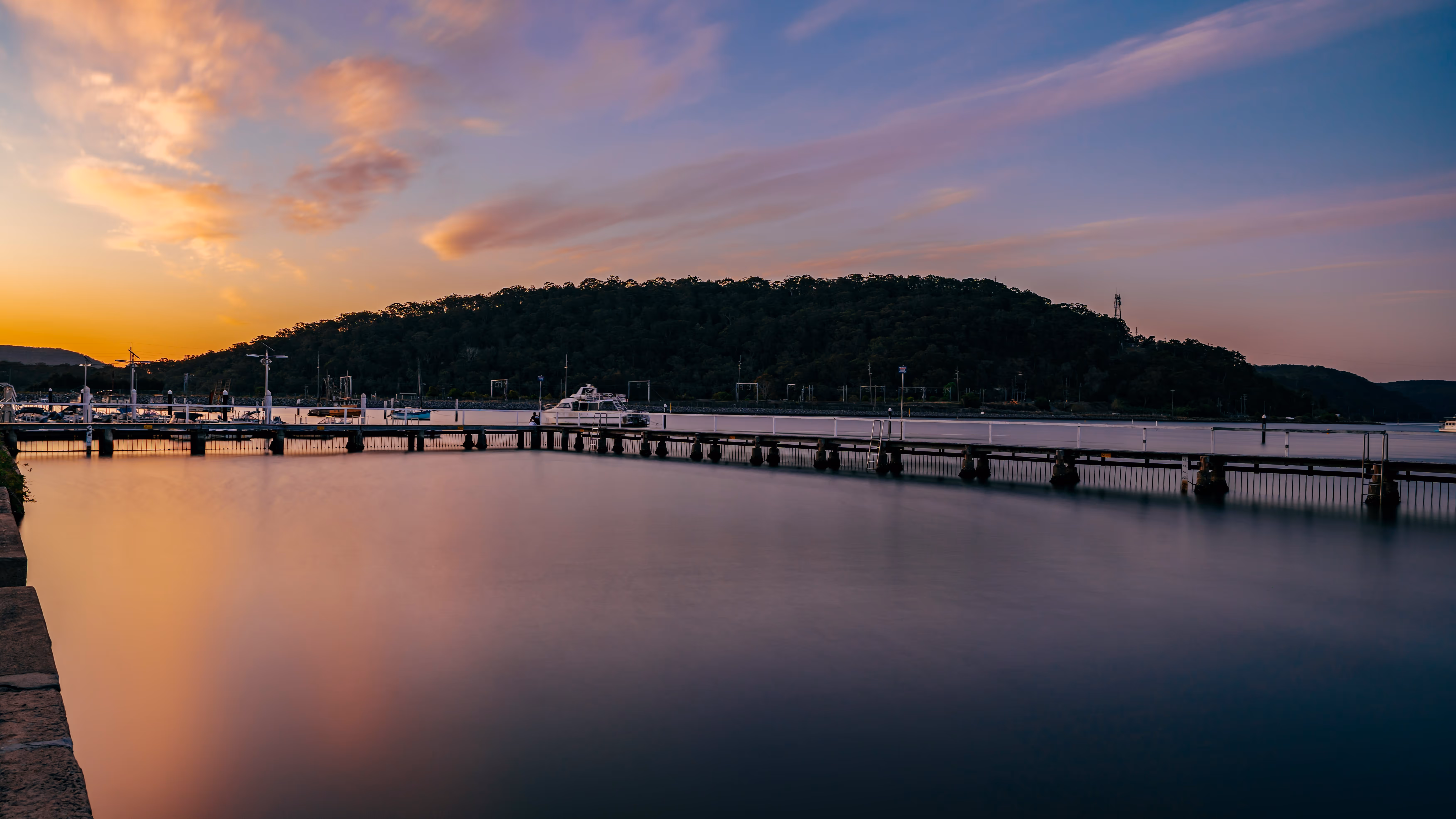 Calm waterfront with a long pier extending into still water and forested hills in the background at sunset.
