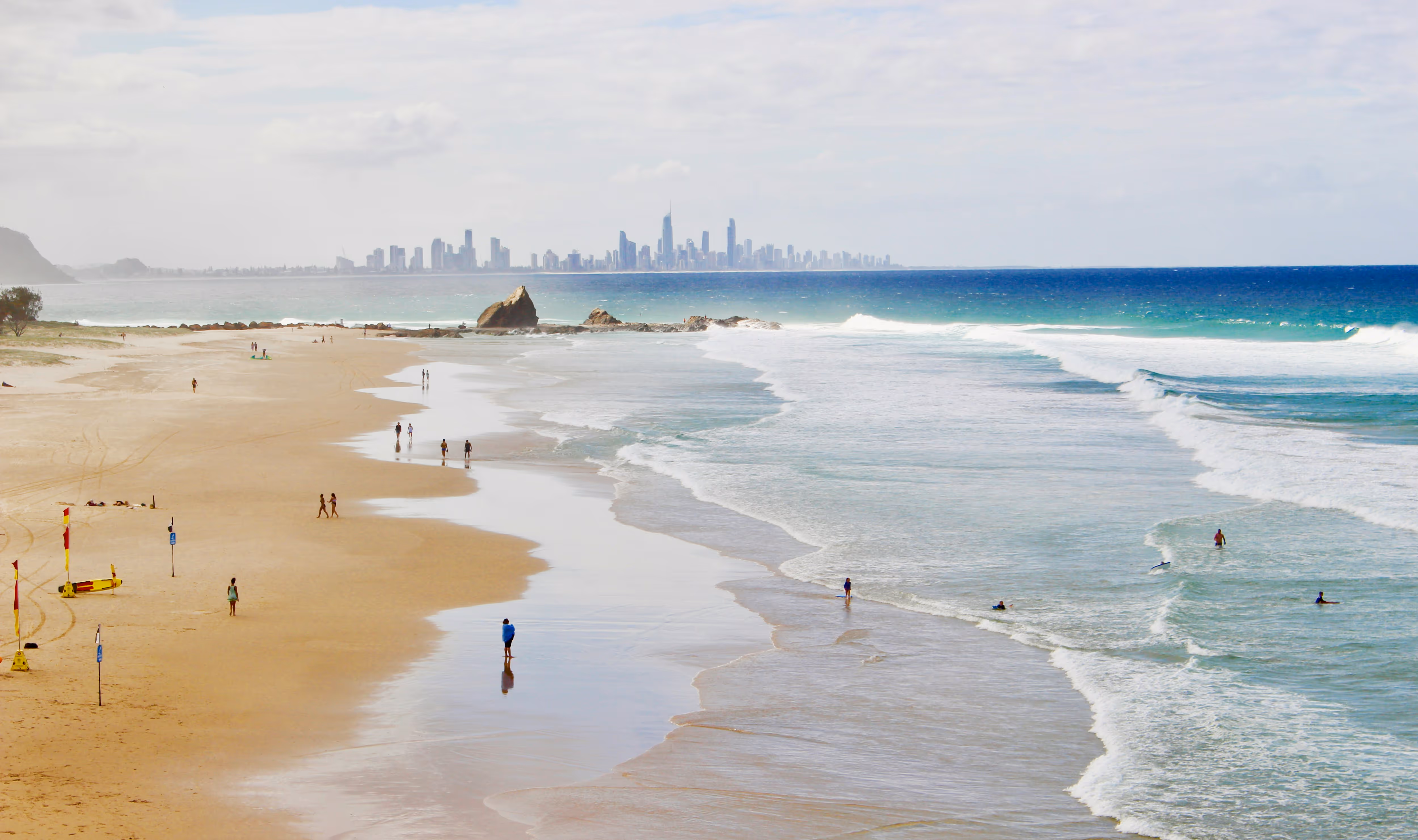 Wide sandy beach with people walking and surfing, gentle waves, and a distant city skyline across the ocean.