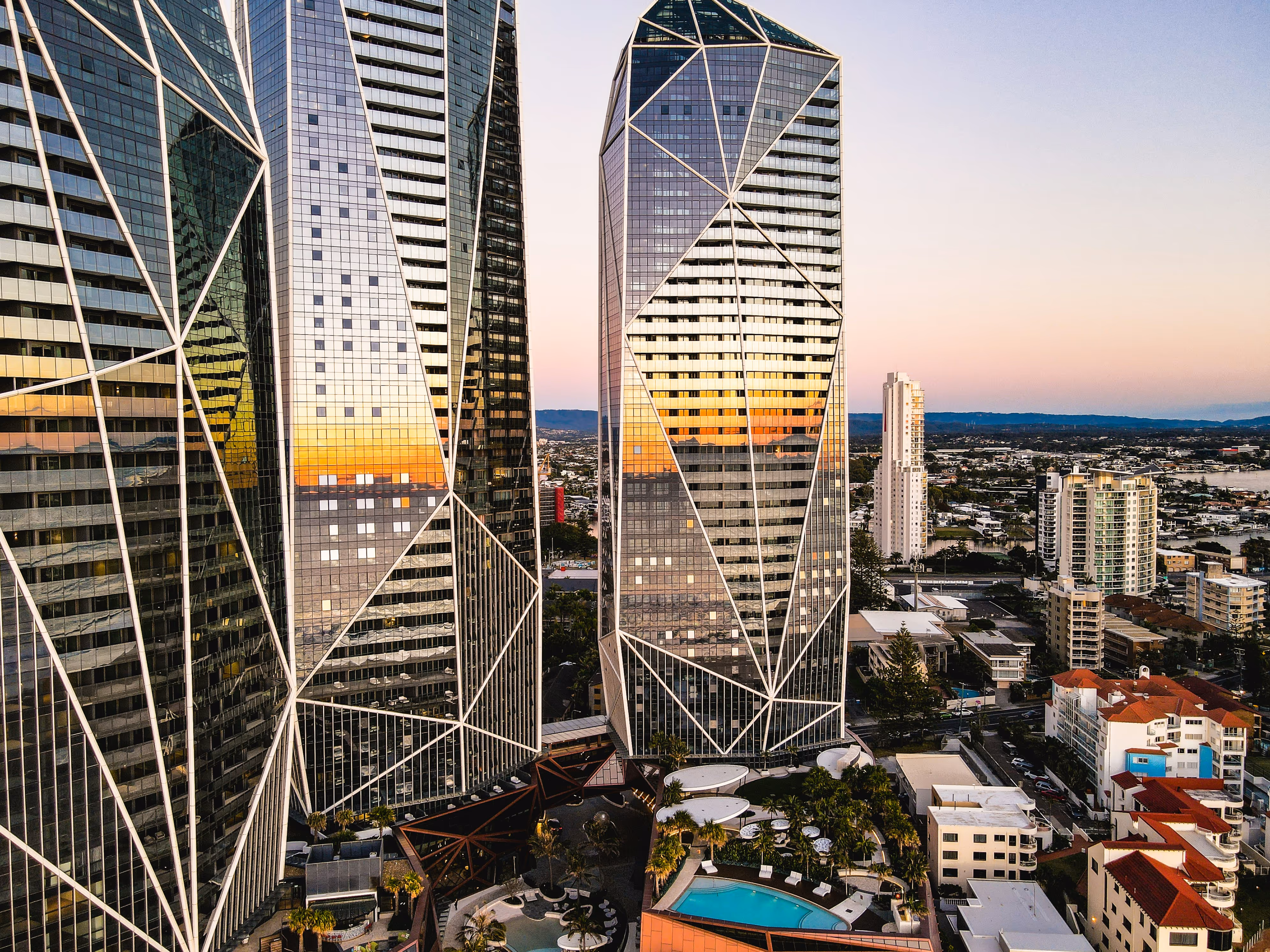 Sunset reflecting on geometric glass skyscrapers above a cityscape with a swimming pool and palm trees below.