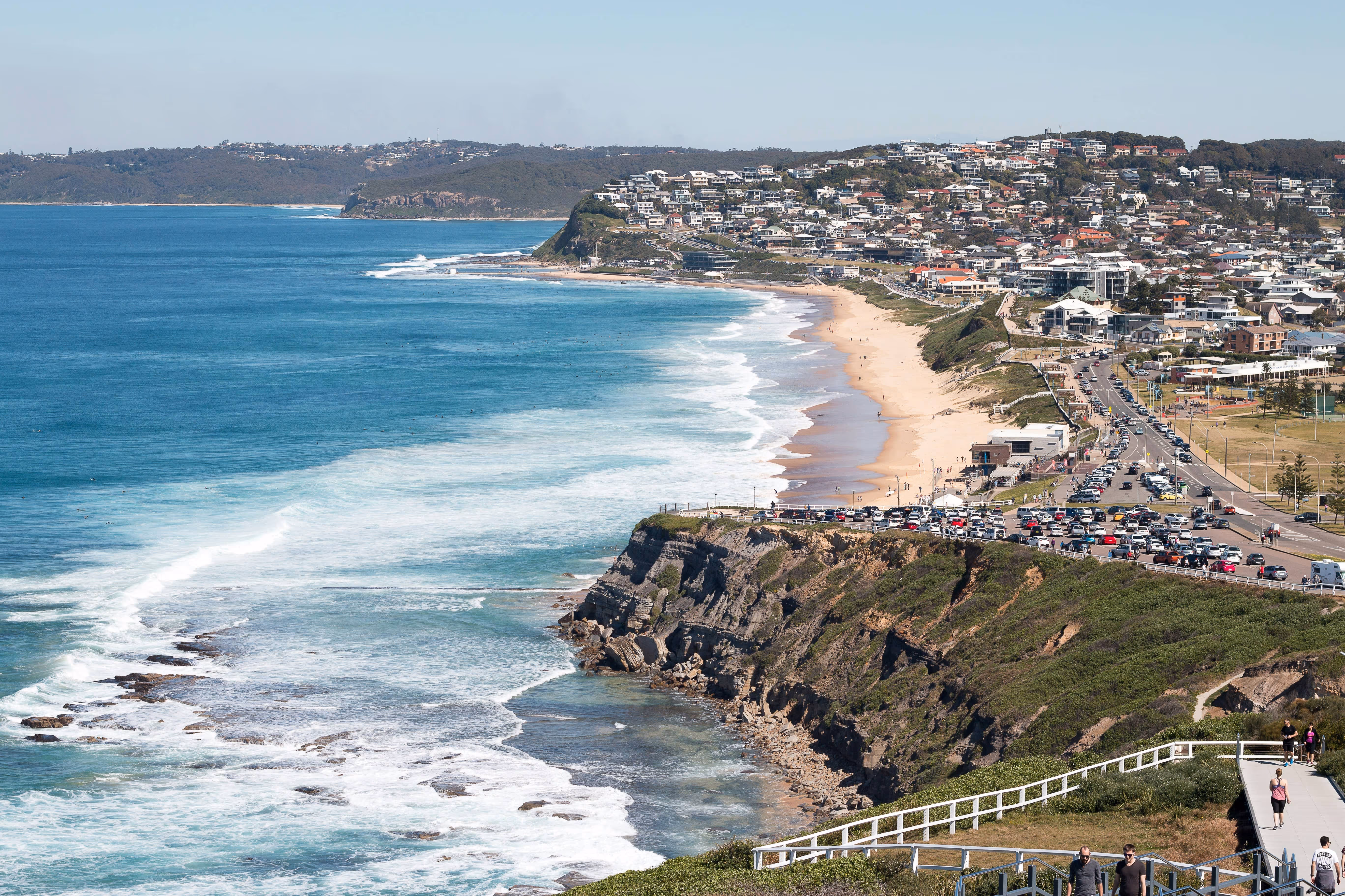 Aerial view of a coastal city with a sandy beach, cliffside, blue ocean waves, and a busy road with parked cars and people walking nearby.