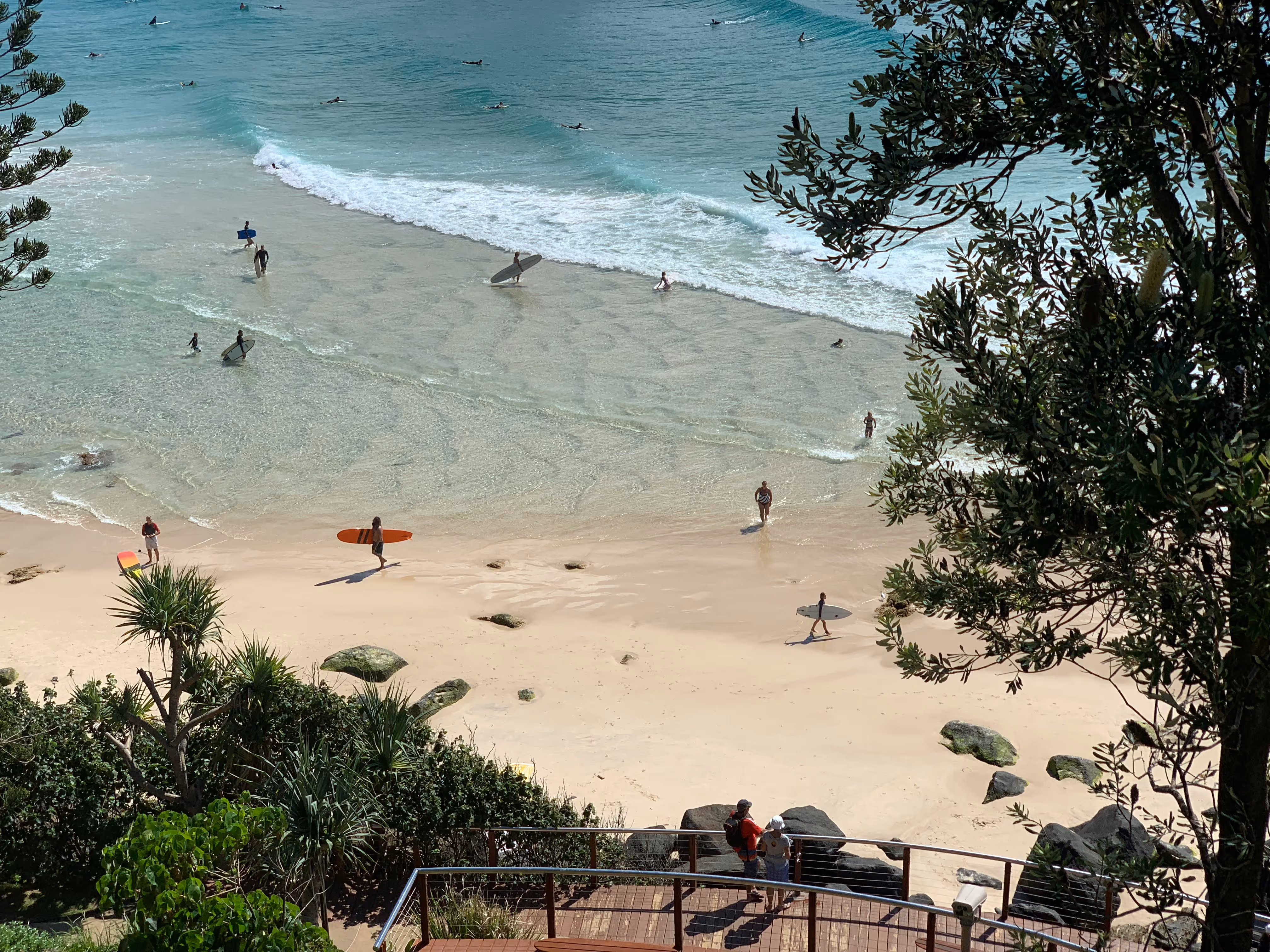 View from above of surfers in wetsuits carrying surfboards on sandy beach and swimming in clear blue water with small waves.