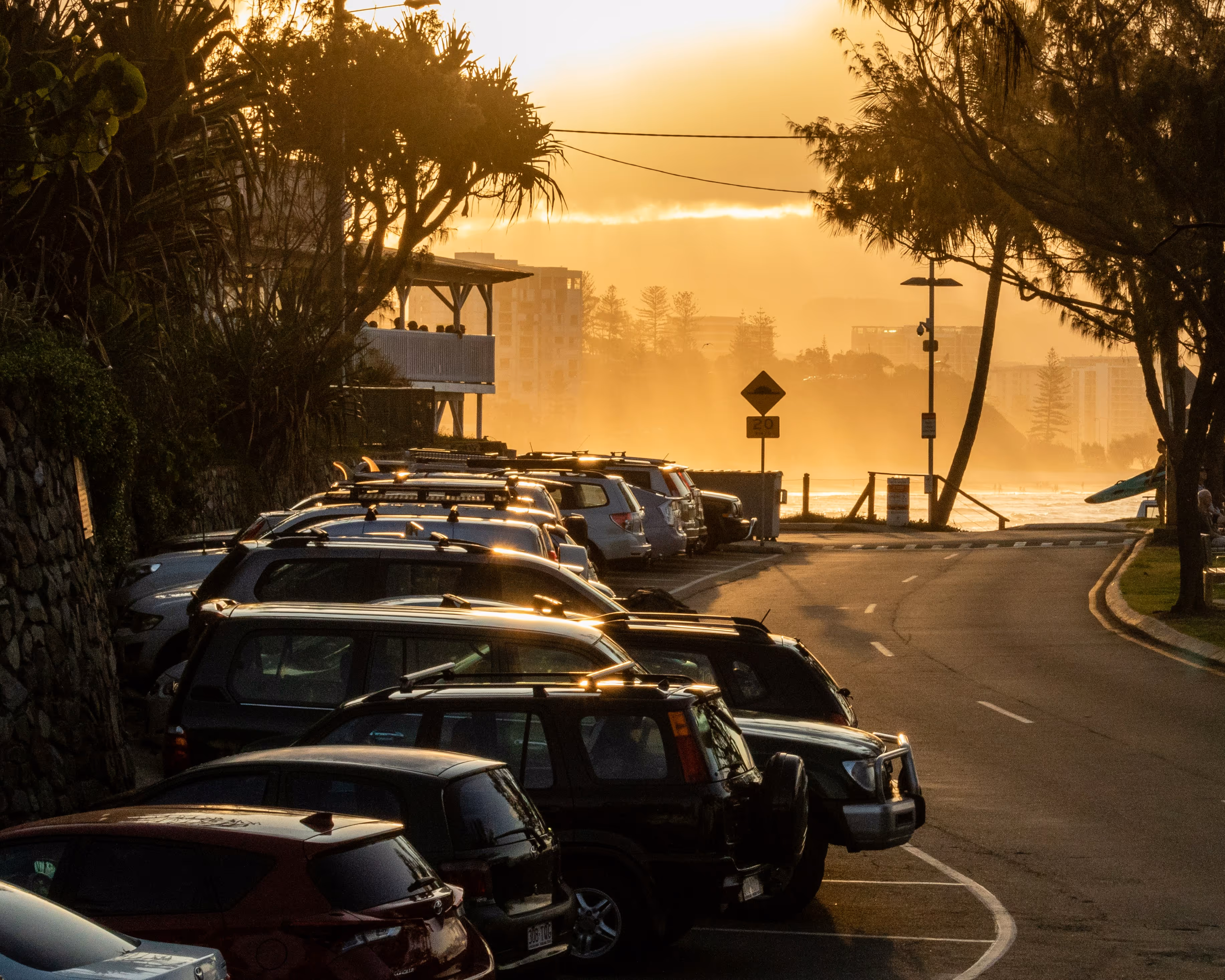 Sunset view of parked cars along a coastal road with trees and buildings in the background.
