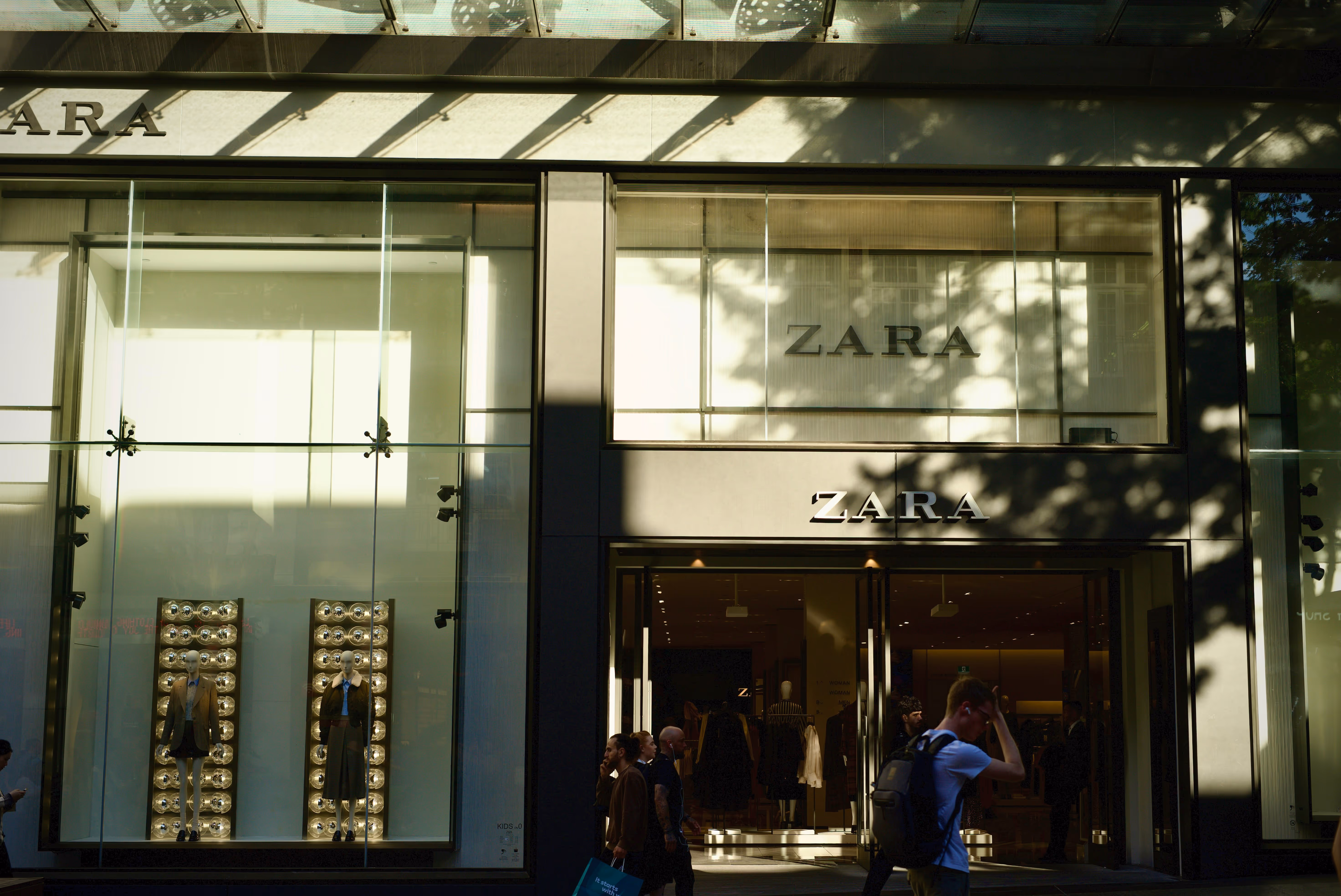 Exterior of Zara store with large glass windows displaying mannequins wearing fashionable outfits, and shoppers walking by.