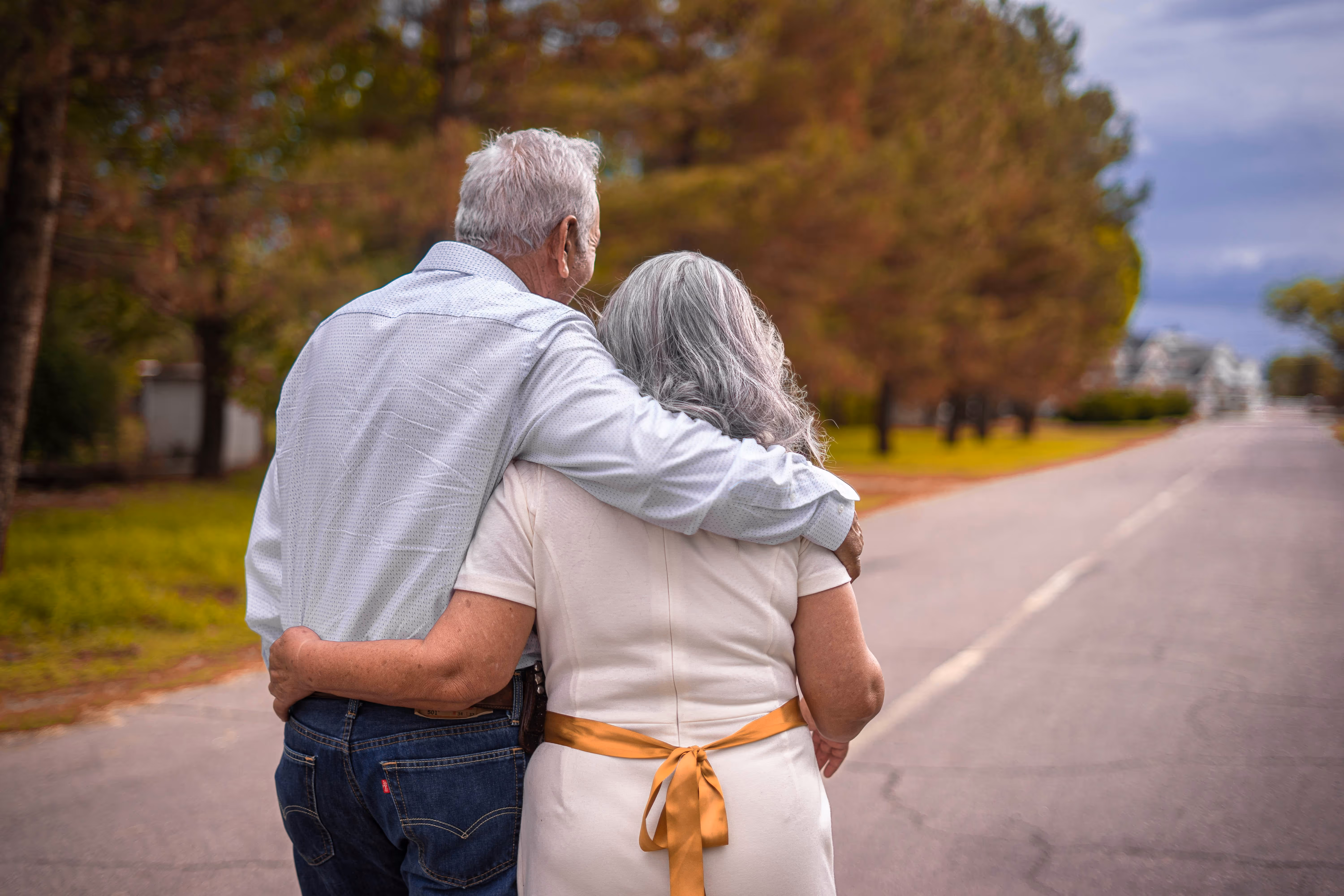 Elderly couple walking arm in arm down a tree-lined street with autumn foliage.