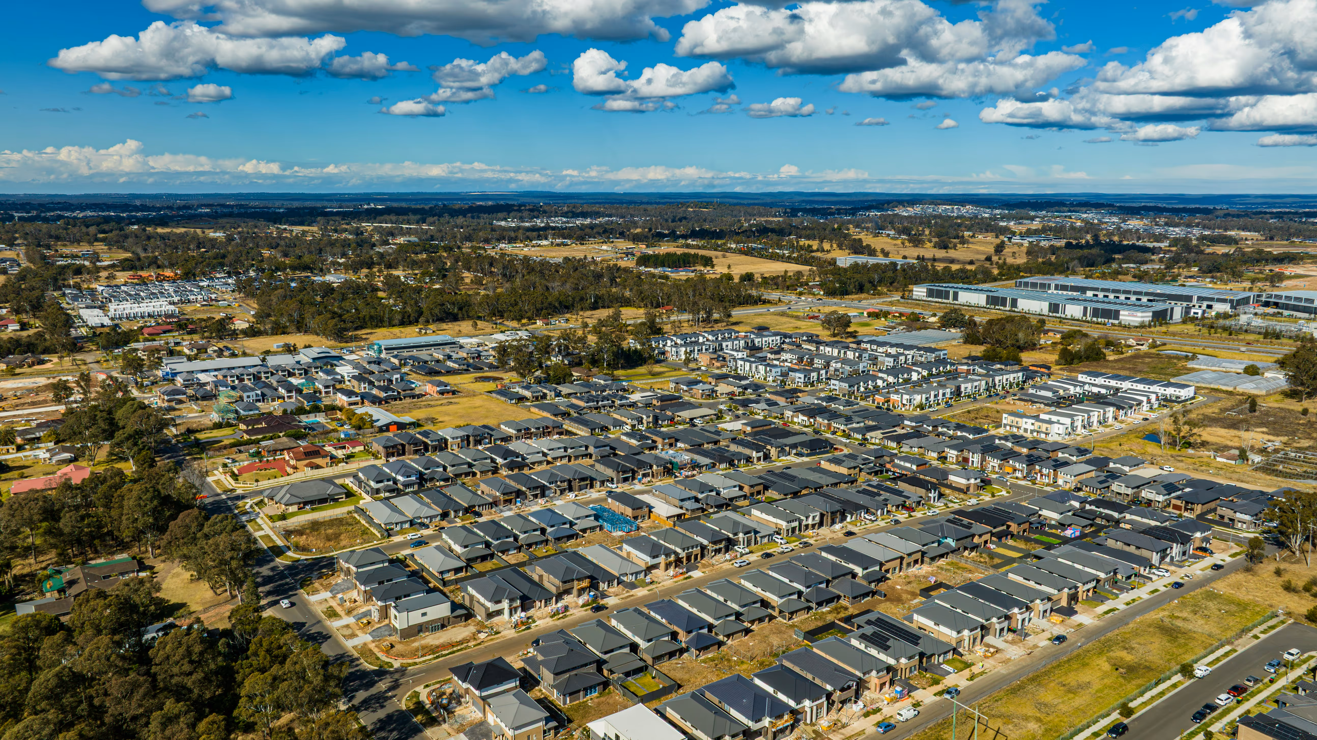 Aerial view of a newly developed Australian neighborhood with modern houses, roads, and surrounding green areas under a partly cloudy sky.
