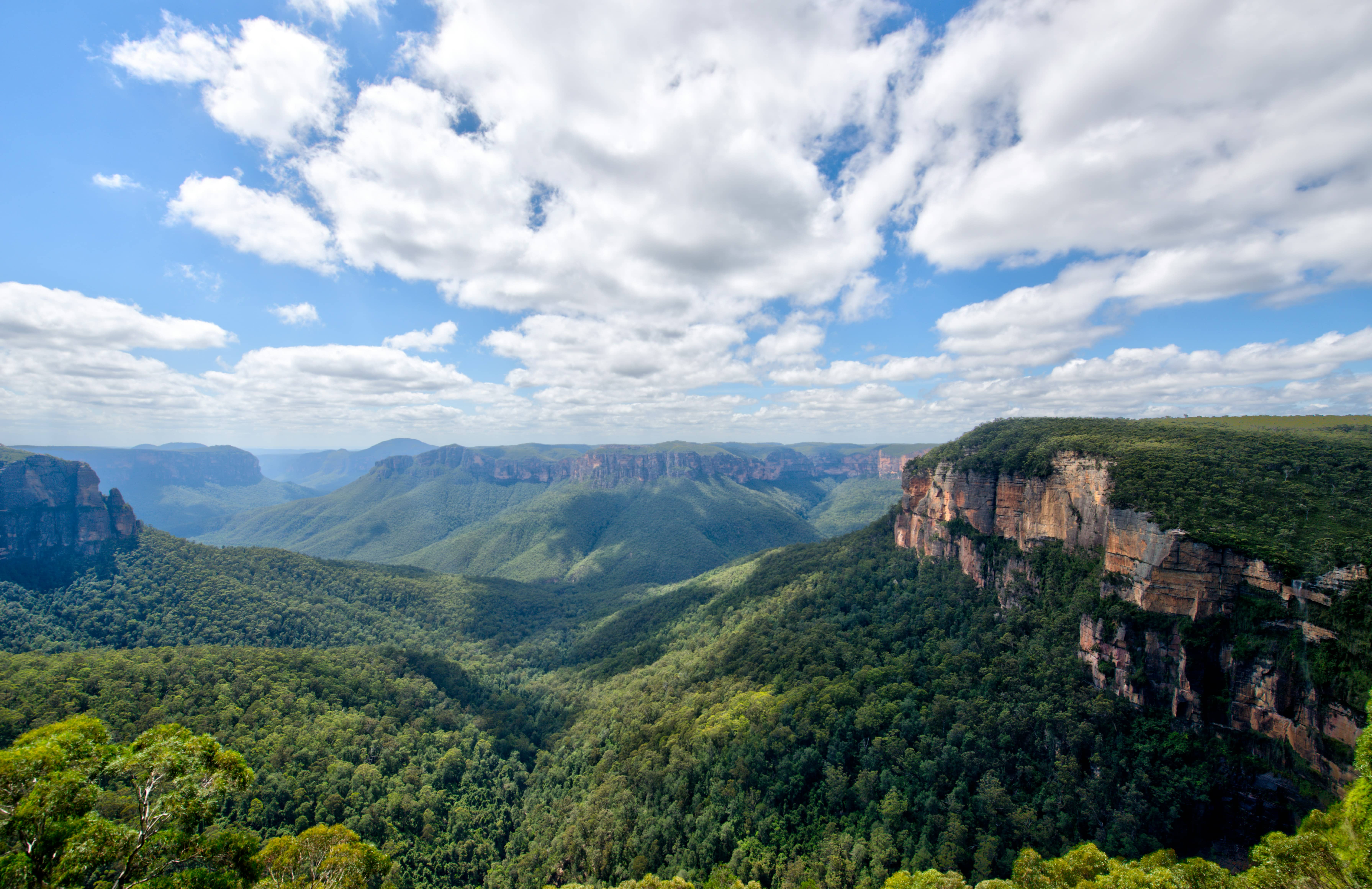 Expansive view of forested mountains under a partly cloudy sky with cliffs on the right side.