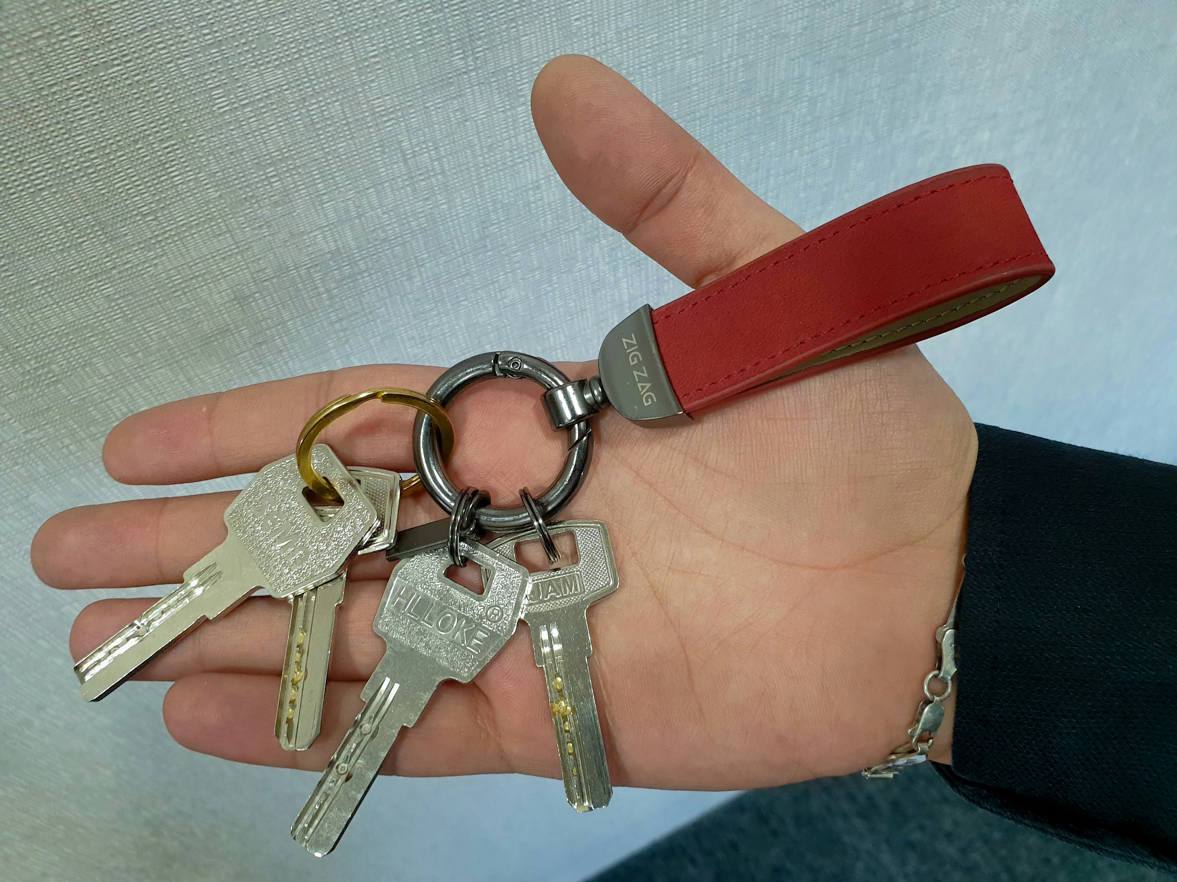 Hand holding a set of metal keys on a keyring with a red leather keychain loop labeled ZIG ZAG.