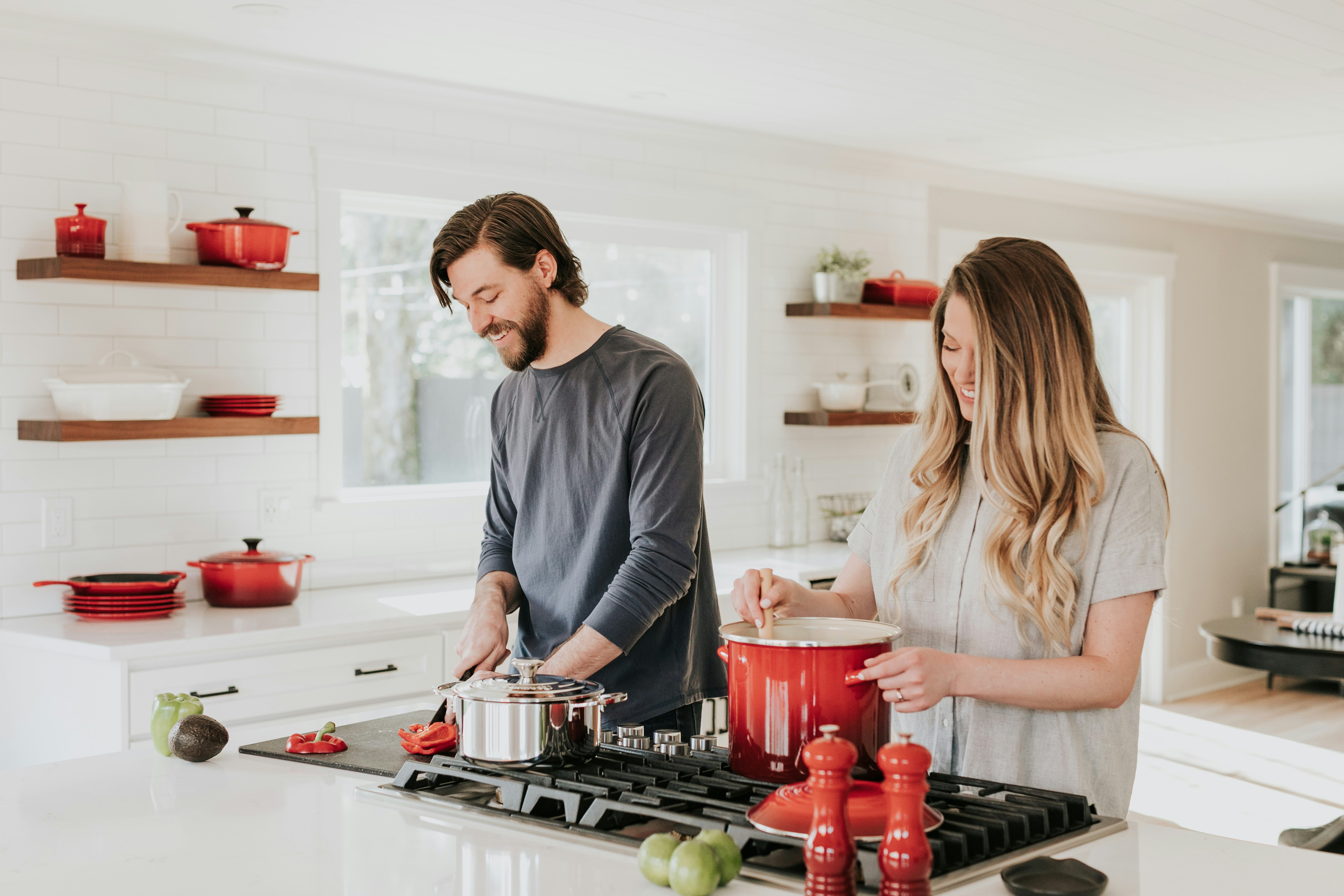 Young couple cooking together in a bright kitchen with red cookware and fresh vegetables on the counter.